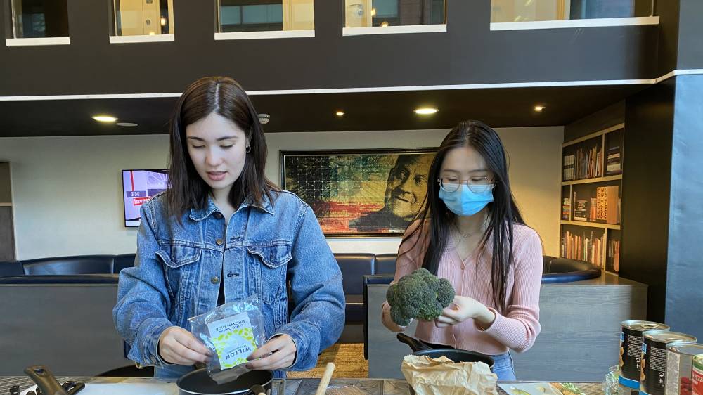 Two people in a kitchen prepare food. One person in a denim jacket holds a package, while the other in a pink sweater and blue mask holds broccoli. Shelves and a framed picture are in the background.