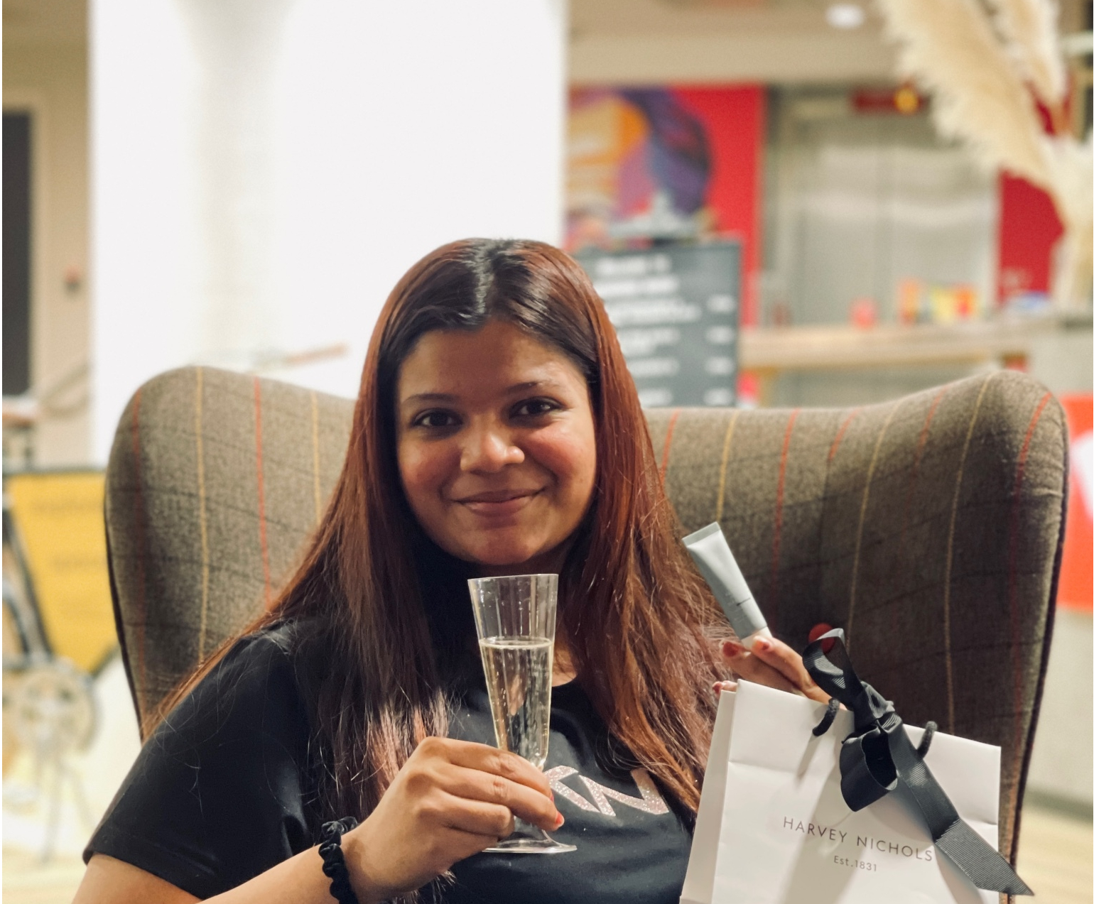 A person with long hair smiles while sitting in a chair at Vita Student, holding a glass of champagne and a shopping bag with a small gift box. The background features a modern interior with subtle lighting, capturing Annette's story of student life and celebration.