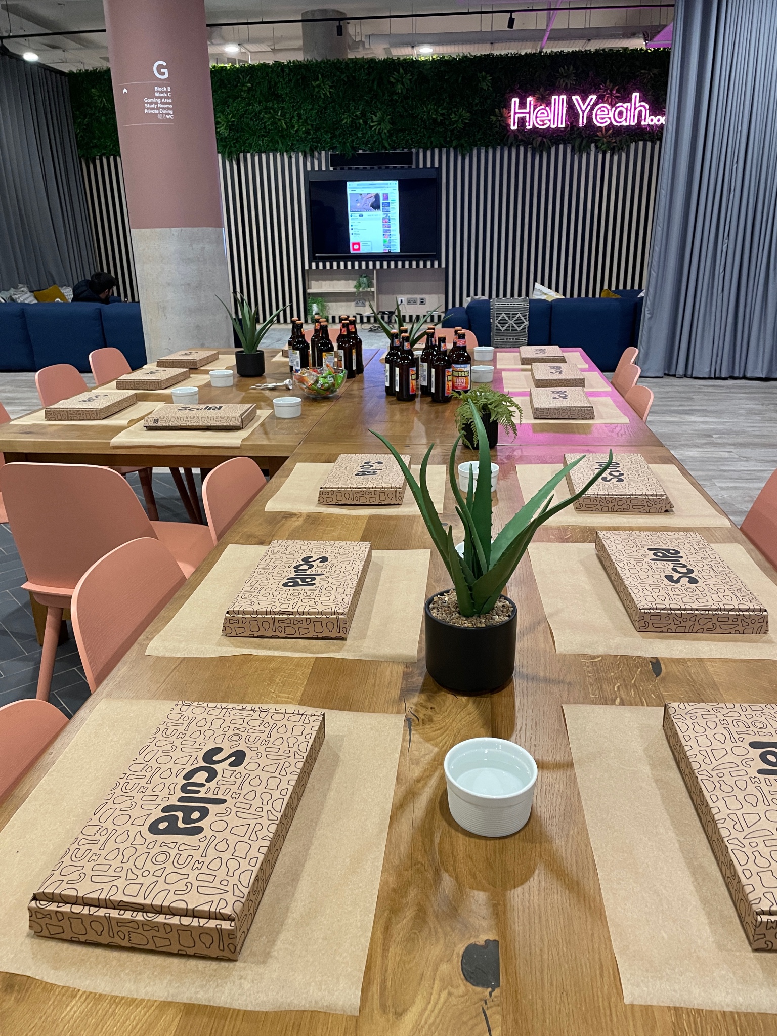 A long wooden table set for a meal with boxed food servings, napkins, and a small potted plant at each place. In the background, an English TV show plays on the wall next to a neon sign reading "Hell Yeah...". The room has modern UK-inspired décor.