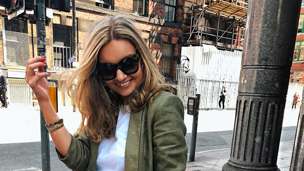 Lydia, with her long blonde hair and sunglasses, smiles while walking outside. She wears a green jacket over a white top. Urban buildings and scaffolding provide an intriguing backdrop, offering insight into the vibrant city life she thoroughly enjoys.
