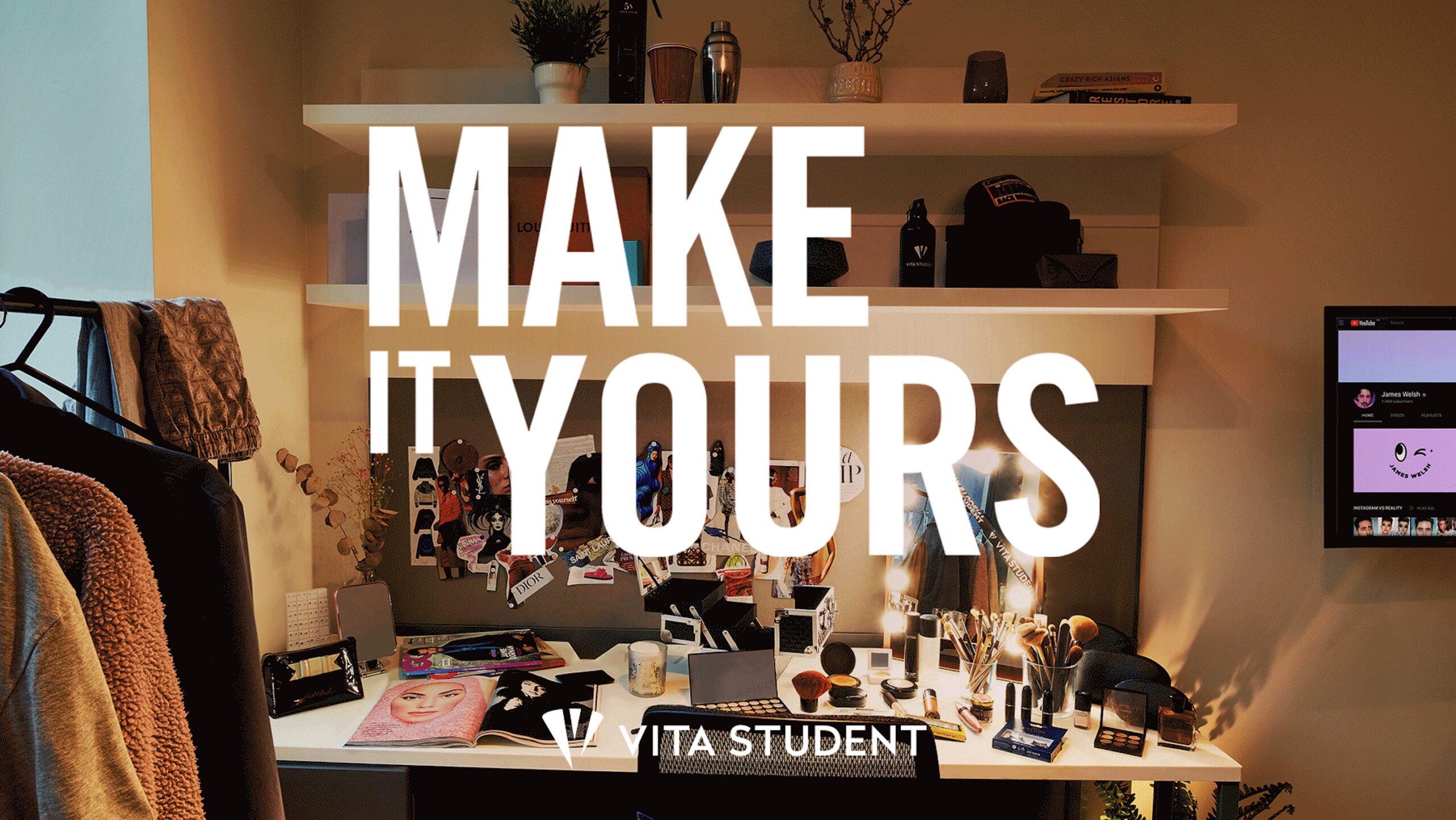 A cluttered desk with makeup, beauty products, and a large mirror with lights. The wall behind has shelves with plants and decorative items. Bold white text overlays the image, saying "MAKE IT YOURS." Clothing hangs on a rack to the side.