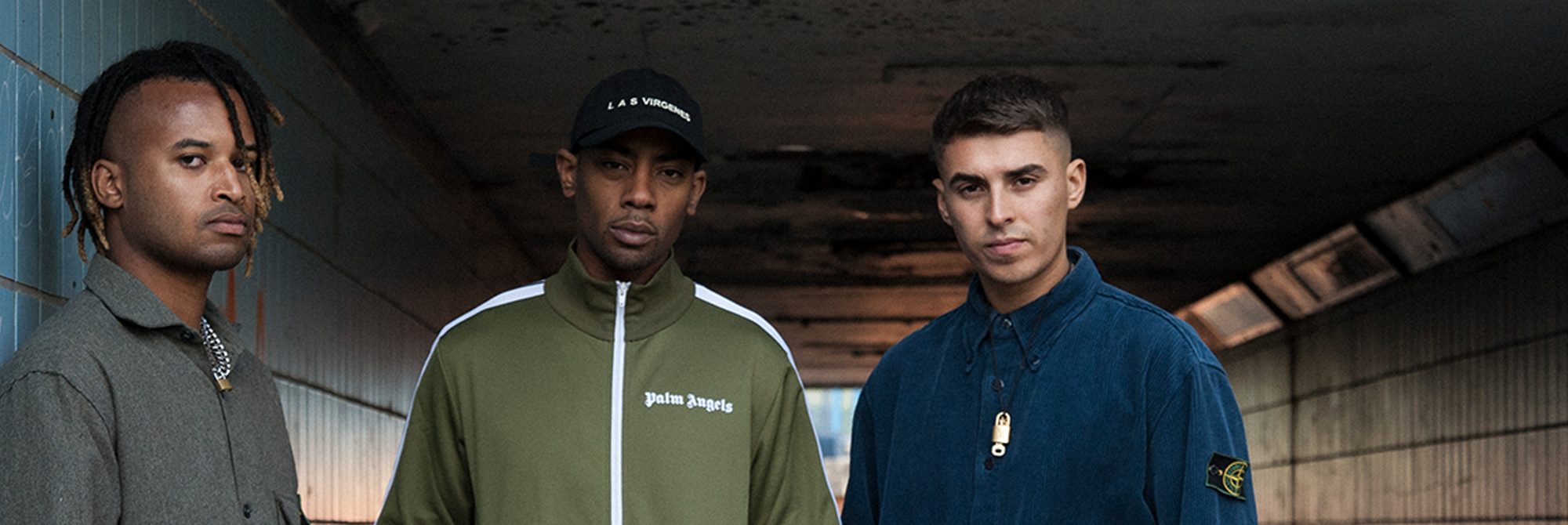 Three men stand in an urban Manchester setting, possibly a tunnel. The man on the left wears a gray shirt, the middle one sports a green jacket with white text reminiscent of Mason Collective's style, and the man on the right wears a blue jacket. The backdrop is concrete with dim lighting.