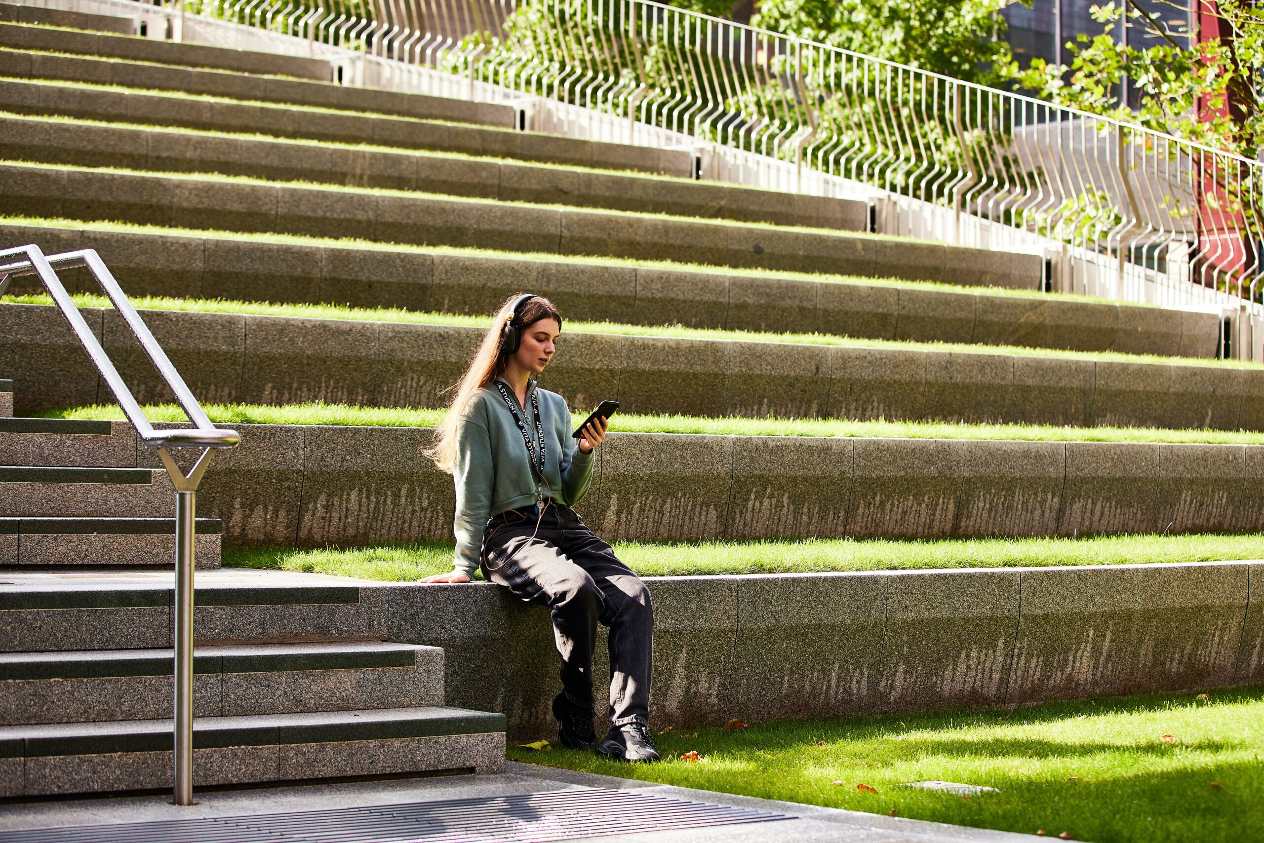 A person with long hair sits on stone steps in an outdoor amphitheater, looking at their phone. They are wearing dark pants and a light green top. Sunlight filters through trees, casting shadows on the steps and grass.