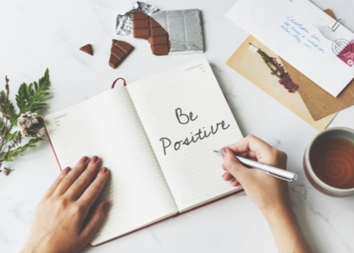On a marble surface, amidst chocolate pieces, a teacup, a small bouquet, and an envelope with a stamp, someone writes "Be Positive" in their notebook. This cozy scene offers an inspiring guide to positivity even in moments of self-isolation.