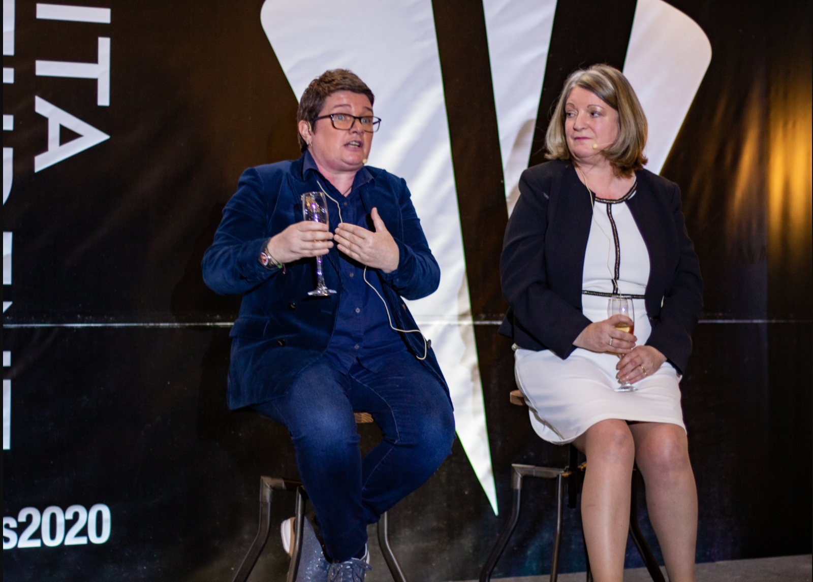 Two people sit on stools in front of a black and white backdrop. The person on the left, wearing glasses, is gesturing while holding a champagne flute—a scene reminiscent of Big Talks. The person on the right, in a white dress and dark blazer, listens intently, reflecting Inspirational Women at their best.