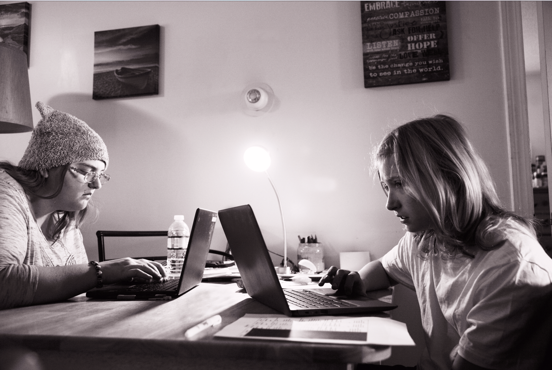 Two people sit at a table, each focused on a laptop. A woman on the left wears a knitted hat, while the person on the right has long hair. A lamp and motivational posters about preparing for challenges are in the background. Papers and a water bottle are on the table, fostering an educational vibe.