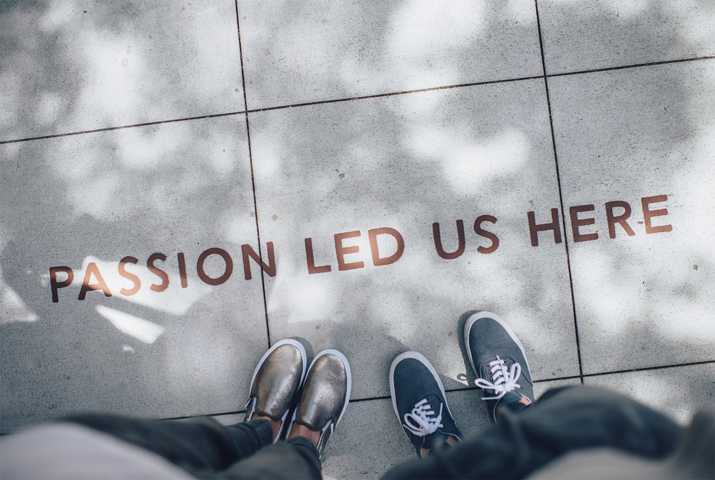 Two people stand on a tiled surface with the words "PASSION LED US HERE" written on it, symbolizing how career skills and dedication can pave the way. The individuals' feet are visible, wearing metallic slip-ons and blue sneakers. Dappled sunlight creates soft shadows on the tiles.