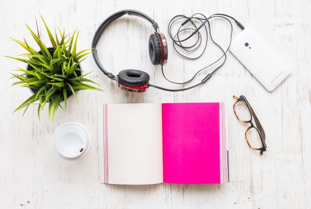 An open book with a bright pink page lies on a white wooden surface, inviting you to read. Nearby are black headphones perfect for listening, a smartphone, glasses, a green potted plant adding life to the scene, and an empty white coffee cup hinting at moments of self-isolation.