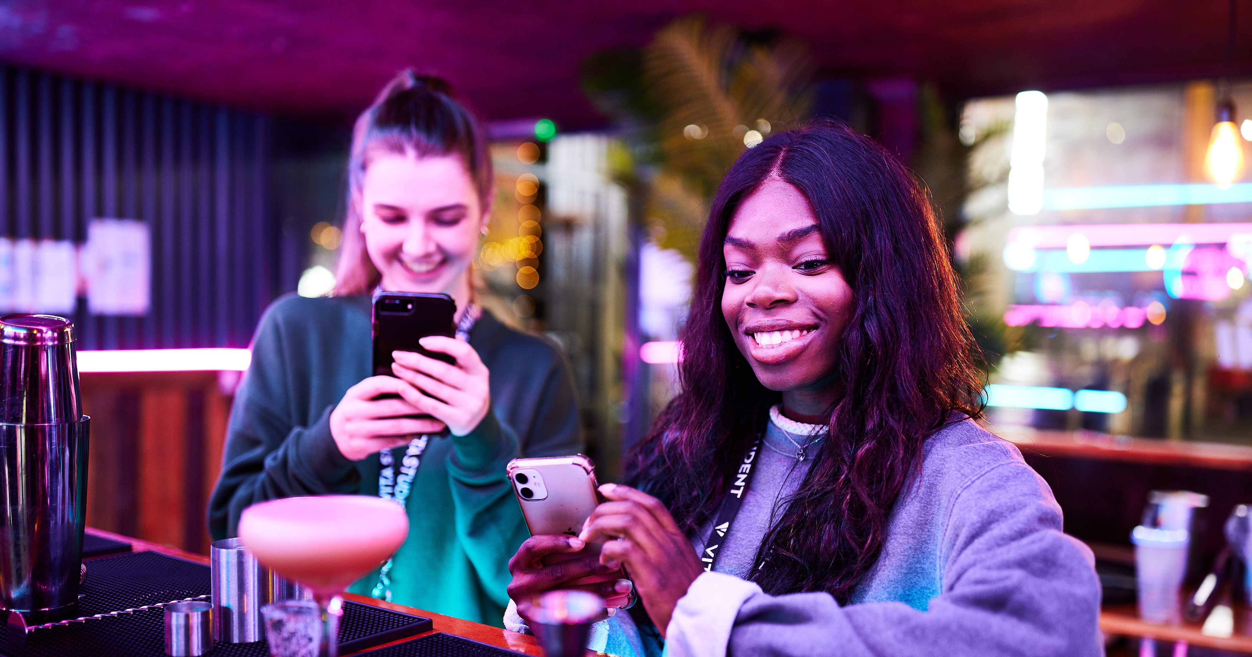 Two women are standing at a bar, smiling while looking at their smartphones. In the brightly lit setting with neon lights, creating a lively ambiance typical of Vita Student's vibrant university life, a pink cocktail sits on the bar in front of them.
