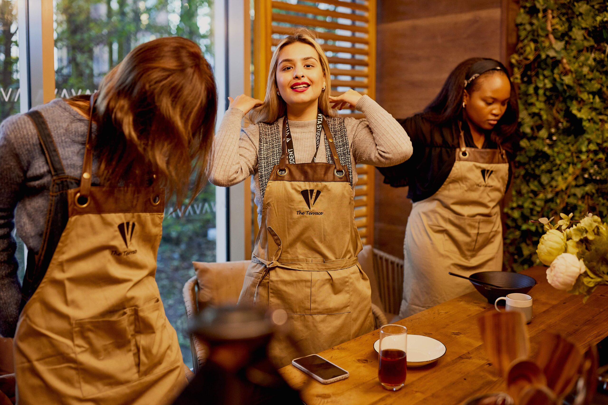 Three people wearing aprons stand at a wooden table, adjusting their straps. A smartphone and drink are on the table, and a green wall is behind them. The group seems engaged in a cooking or workshop setting.