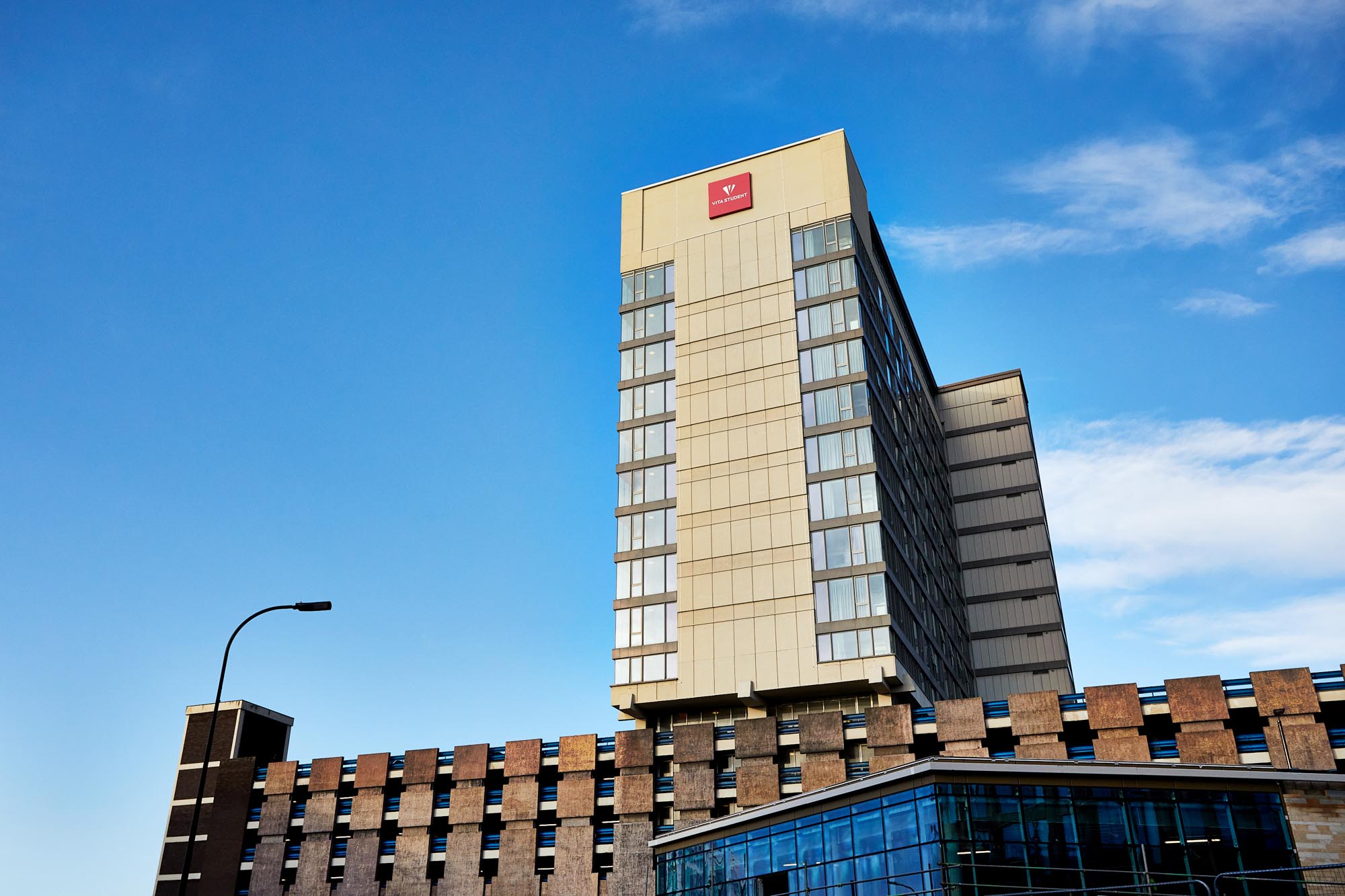 A tall modern hotel building under a blue sky boasts a red "Y" and "W" logo on top. Reflective windows and contrasting brickwork characterize this urban lifestyle hub. In the foreground, a blue glass front hints at chic student living, with a streetlight visible to the left.