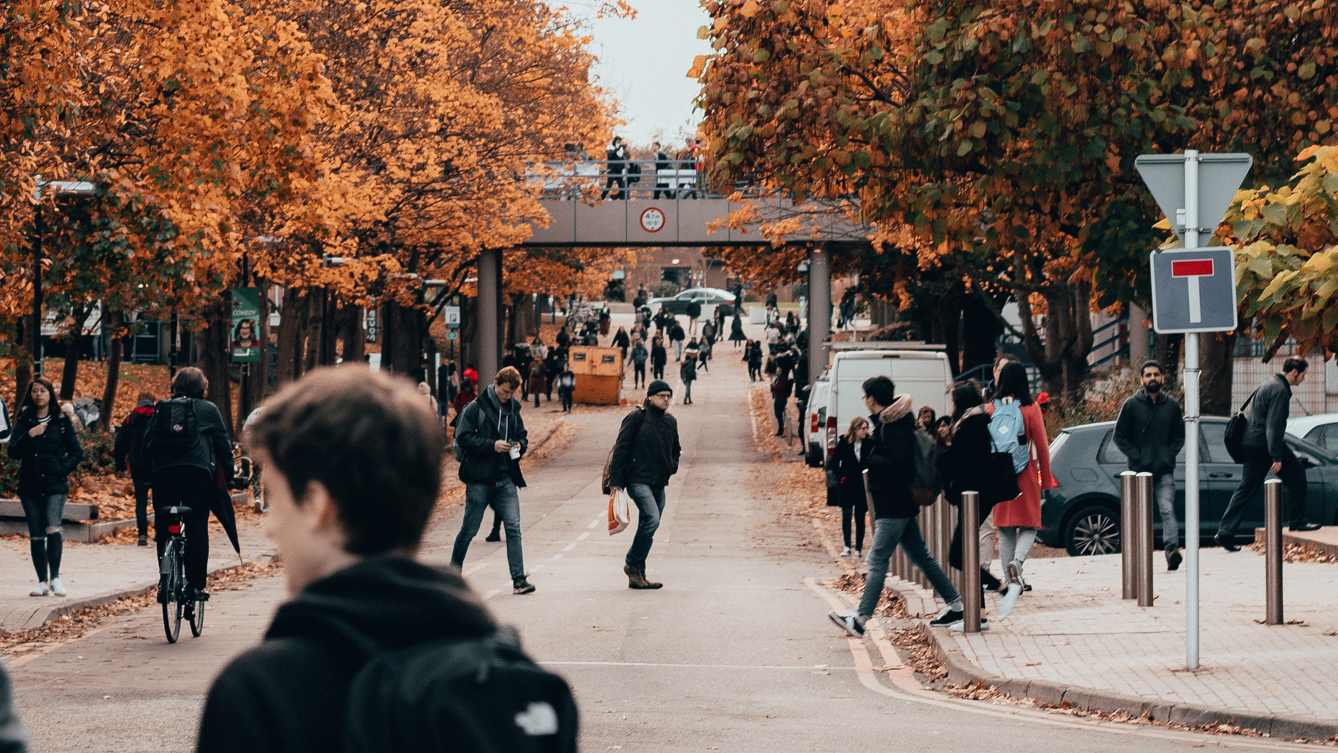 A bustling street scene in autumn with people walking and cycling near Warwick. Trees lining the street display vibrant orange leaves. A pedestrian bridge in the background has people crossing over. The atmosphere is lively and urban, inviting you to explore every corner of this vibrant setting.