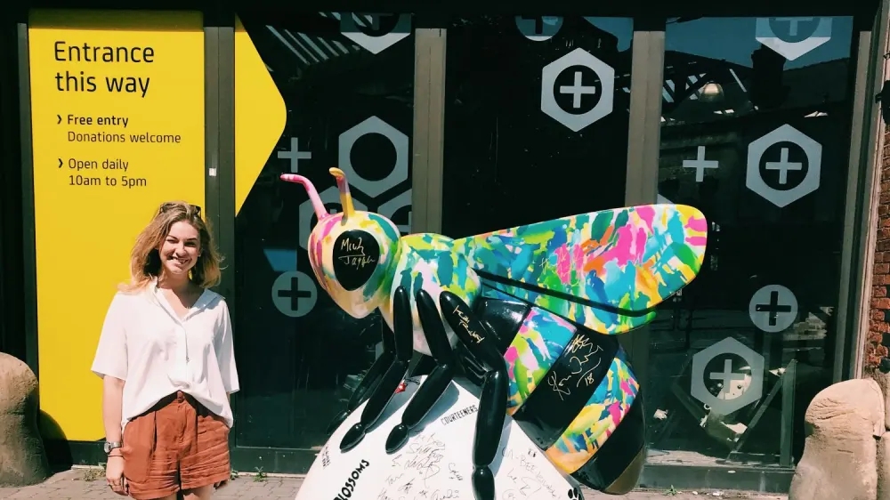 A woman stands smiling next to a colorful bee sculpture covered in various signatures by a Manchester artist. Behind her is a yellow sign indicating entrance directions and general information.