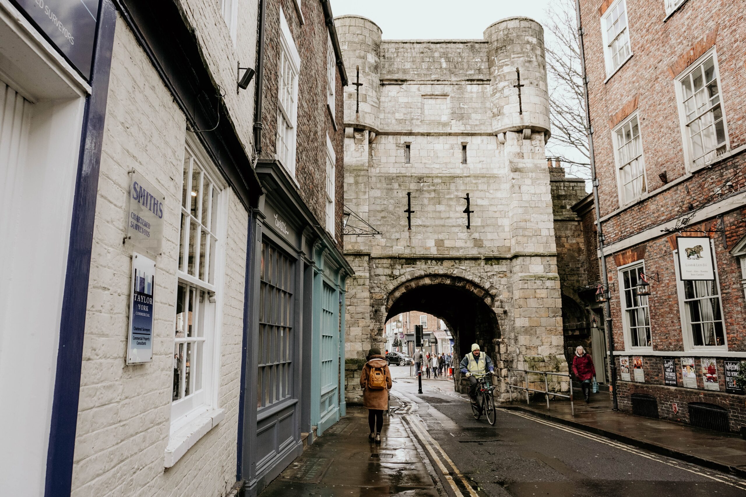 A narrow, wet street in York leads to a stone archway. A person wearing an orange jacket walks alongside a cyclist. The street is lined with old brick and stone buildings, inviting you to explore York's historic charm. The sky is overcast.