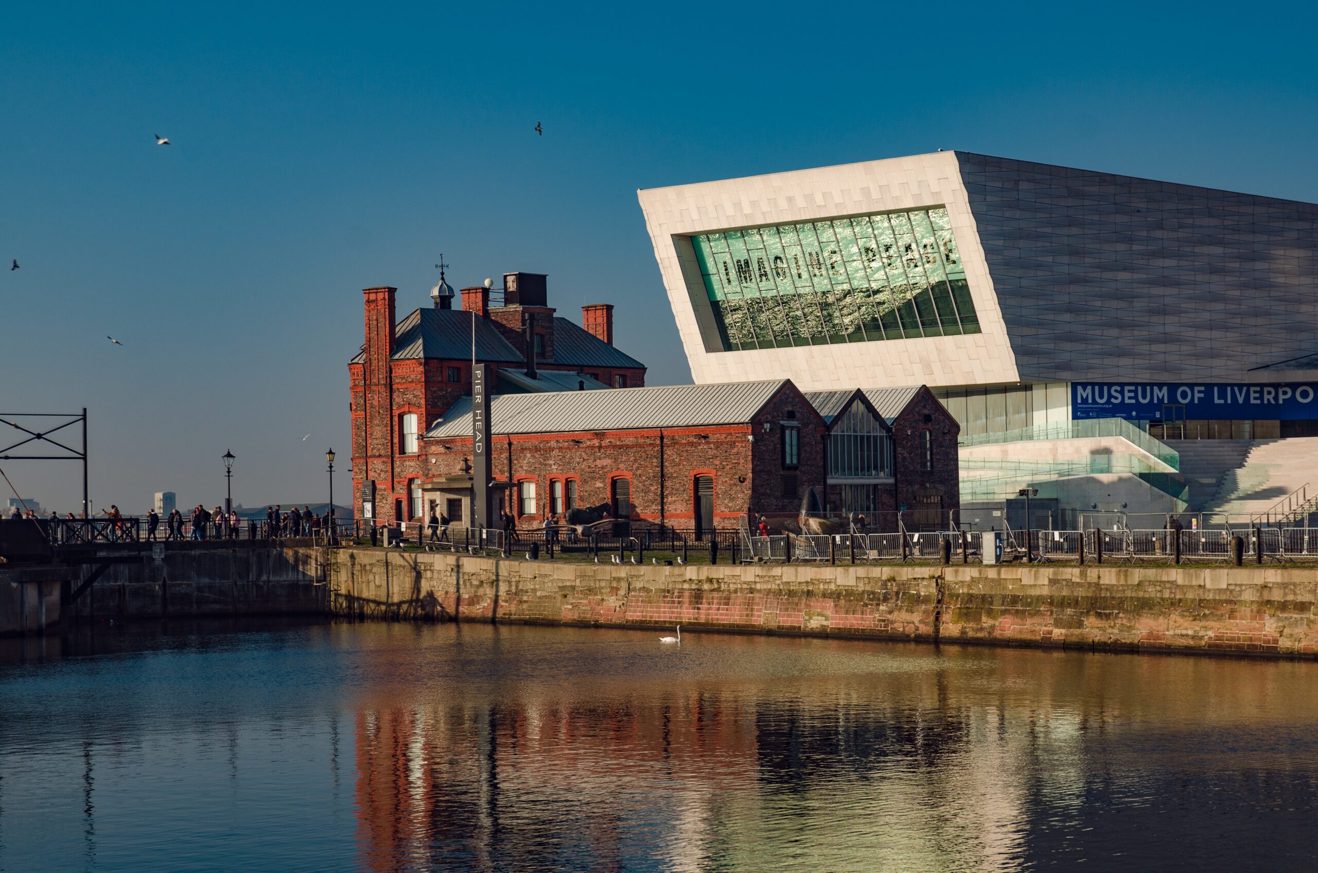 View of two buildings by the water in Liverpool, one with a modern angular design and the other showcasing traditional red brick architecture. The water reflects the structures, a clear blue sky is overhead, and students explore things to do while birds float in the background.