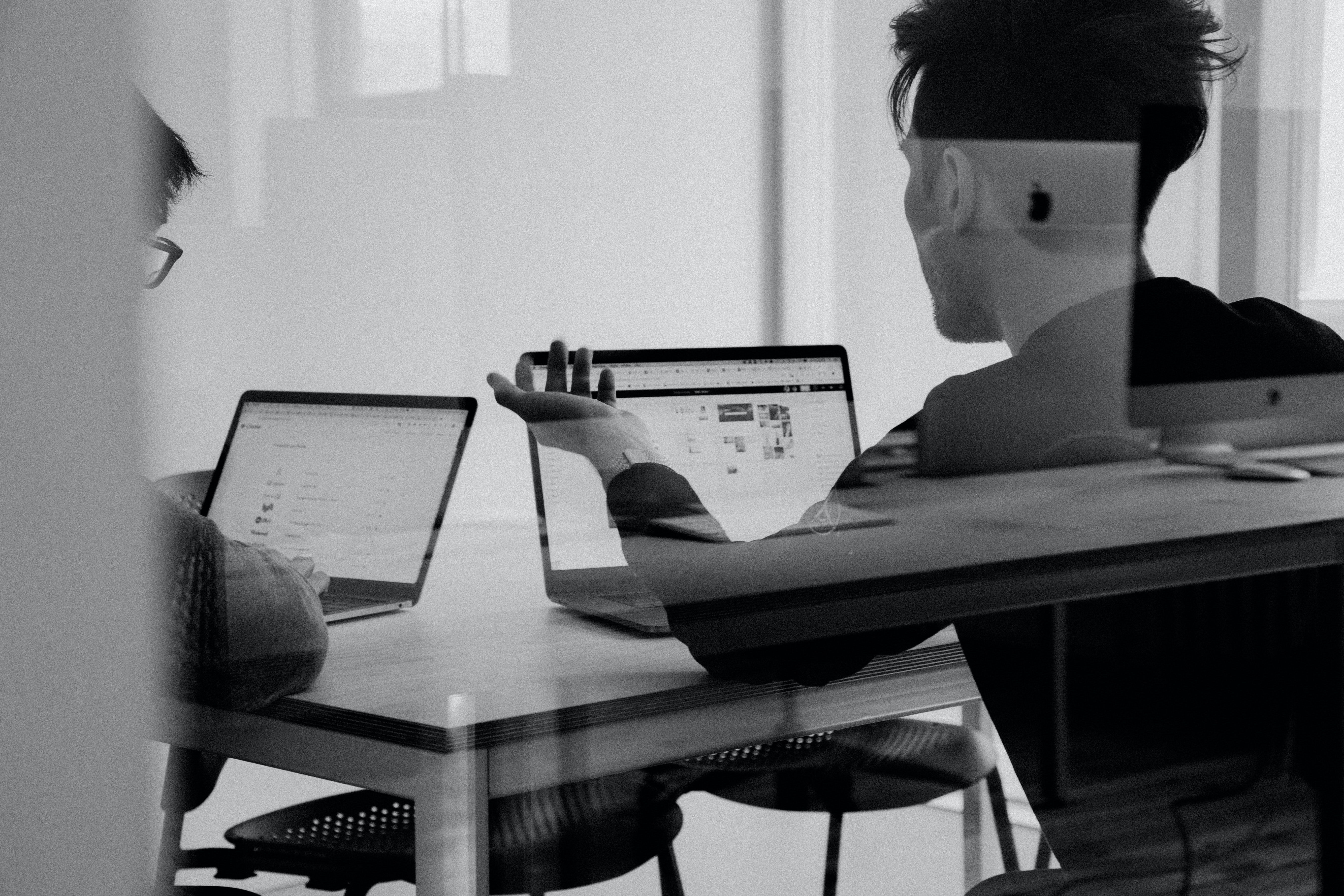 Two people sit at a table with laptops open in a Vita Student lounge. One person gestures toward their laptop screen, suggesting a discussion or collaboration on start-up ideas. The scene is captured through a glass partition, adding a reflective effect.