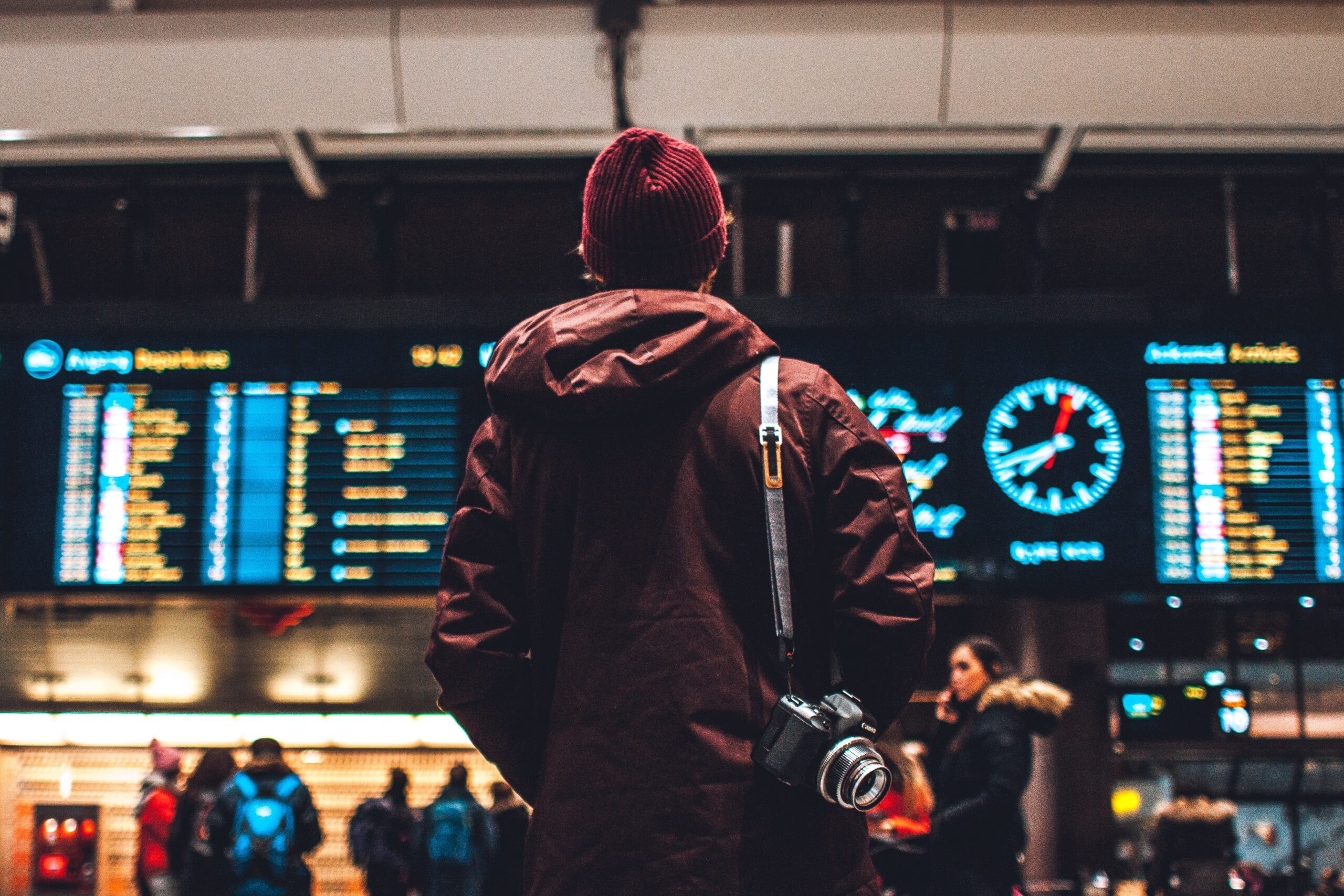 A person in a brown coat and red beanie stands facing an airport departure board, embarking on their journey. With a camera hanging from their shoulder, the board displays flight details. People and luggage bustle nearby, perhaps some heading to sunny Cyprus or back to Vita Student life.