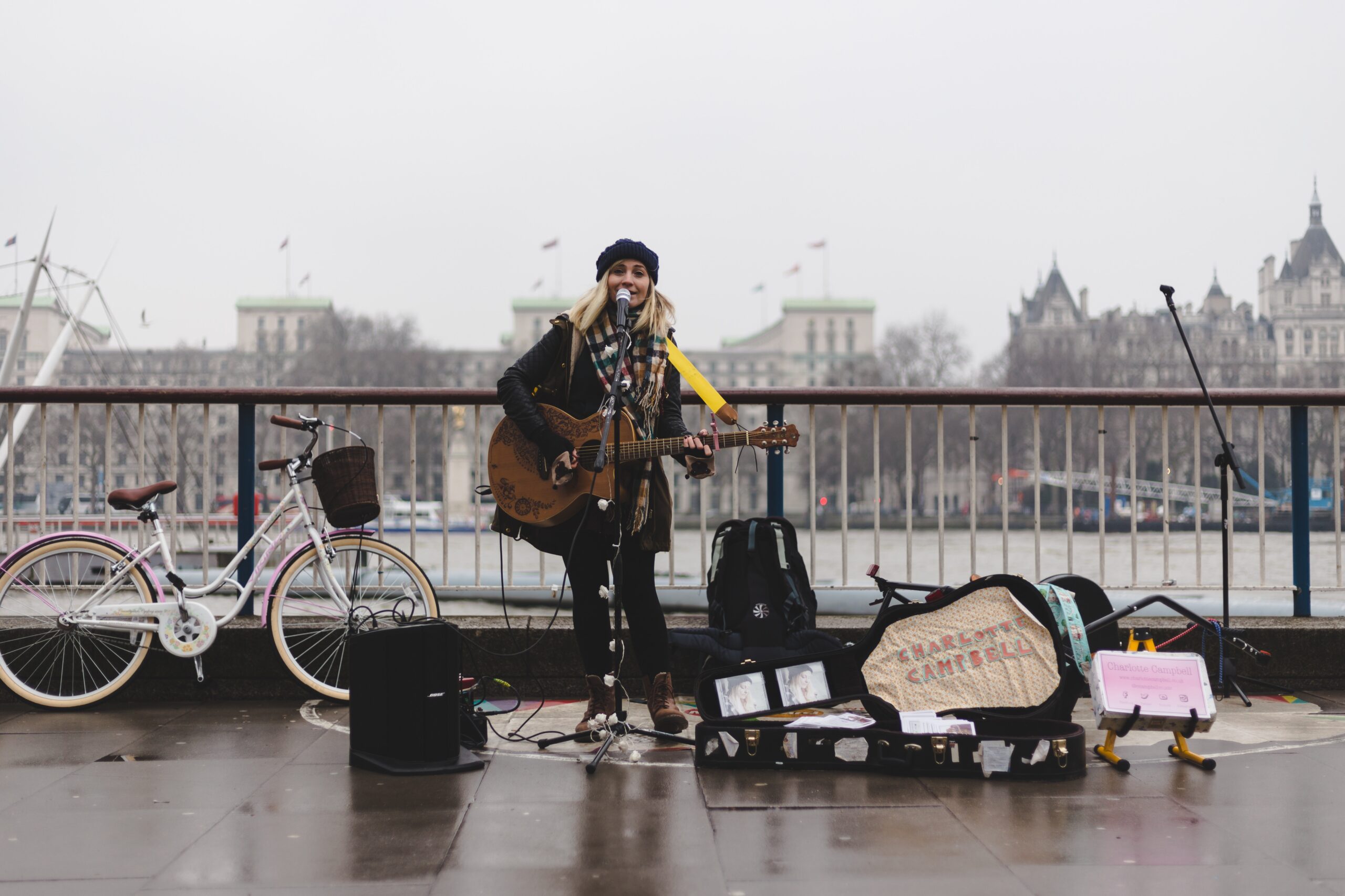 Street performer sings and plays guitar on a riverside promenade, hoping to gather some funding. A microphone stands in front, with an amplifier and open guitar case filled with items. A bicycle is parked nearby. Buildings and a Ferris wheel are visible in the foggy background.