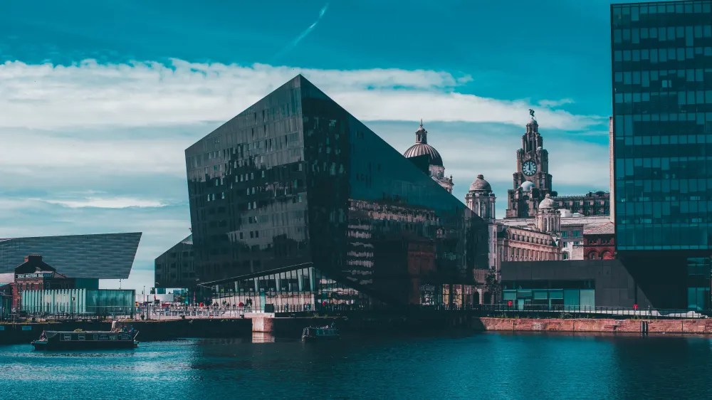 The modern black glass building by the waterfront, offering luxury student accommodation, is framed by historic architecture in the background, including a clock tower. The sky is clear with some clouds, and the water is calm.