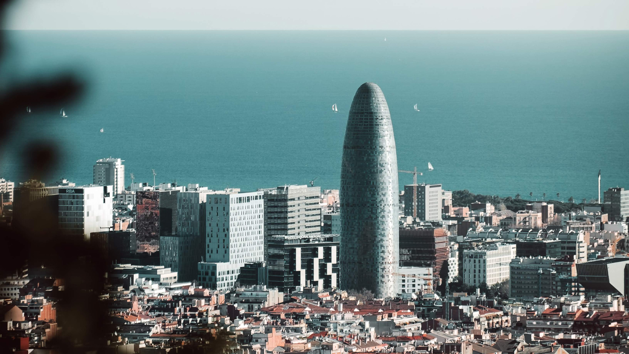 Cityscape of Barcelona featuring the Torre Glòries skyscraper. The cylindrical building stands prominently against a backdrop of the Mediterranean Sea with small sailboats visible, alongside modern and traditional structures near Vita Student Poblenou accommodation in this lively neighborhood.
