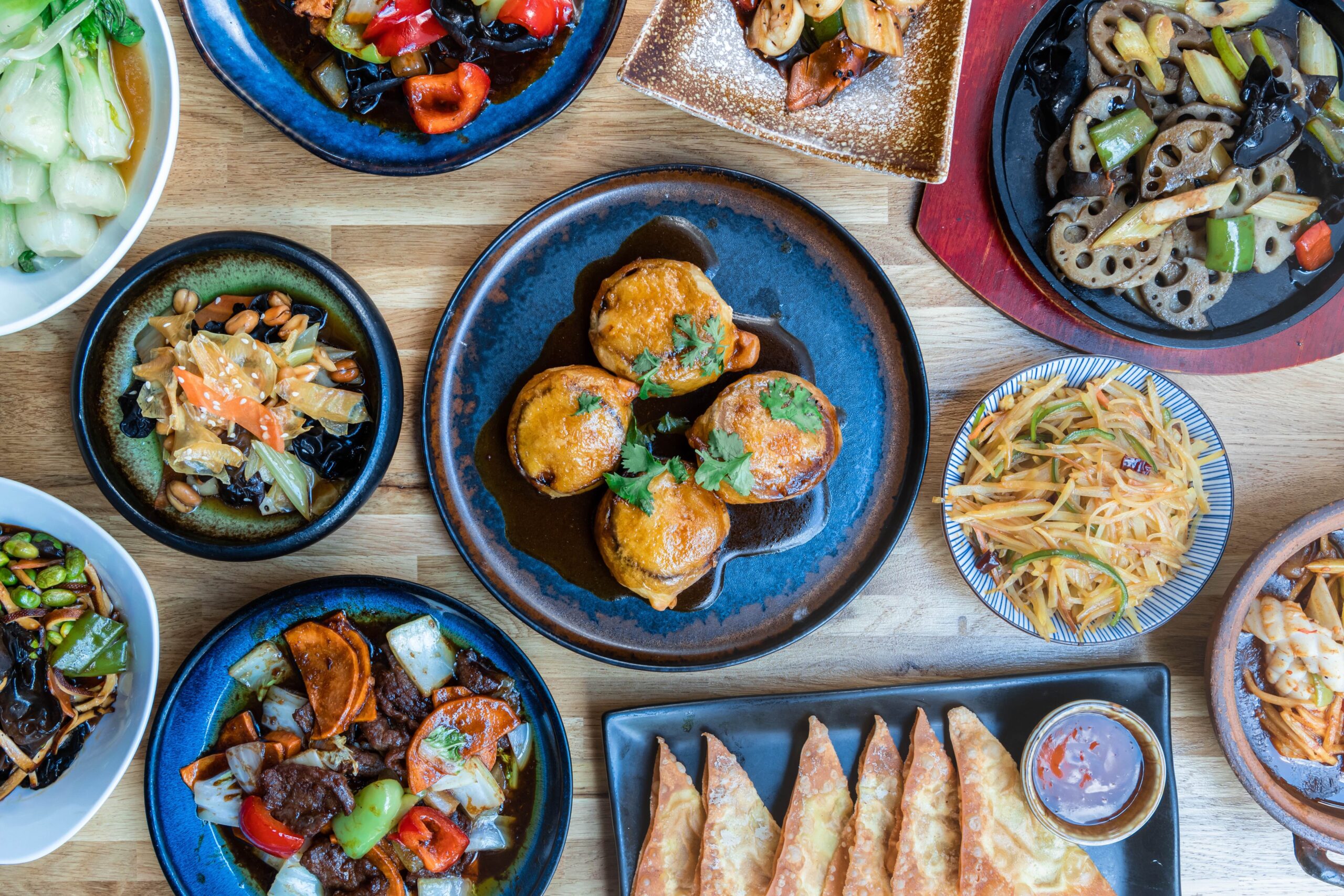 A colorful display of various Asian dishes at a Thai restaurant in Exeter, set on a wooden table. Bowls and plates brim with vegetables, meats, crispy fried items, noodles, and dipping sauces, all artfully arranged for a vibrant and appetizing presentation.