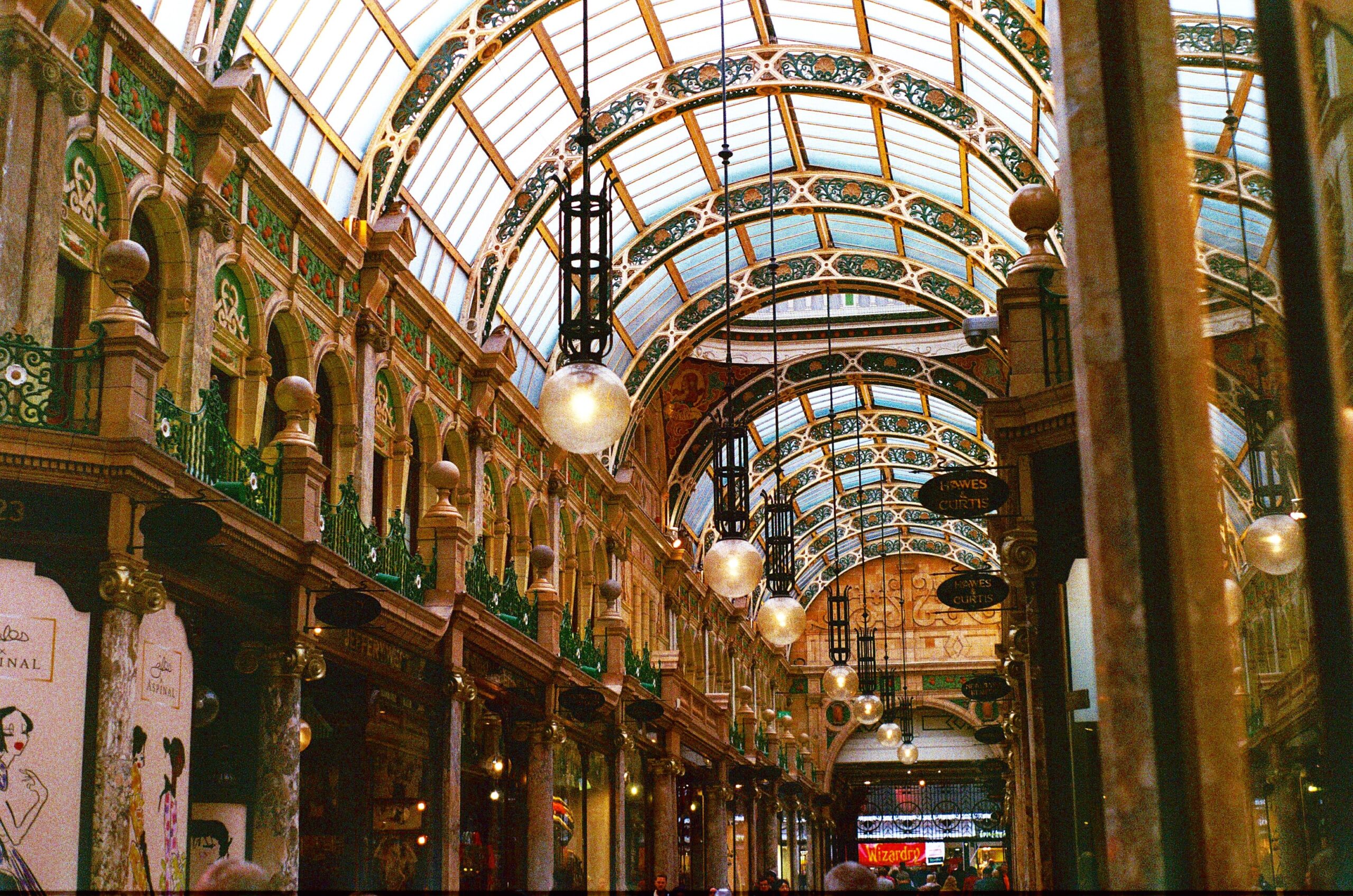 Ornate shopping arcade in Leeds with a glass-paneled vaulted ceiling, decorative arches, and hanging globe lights. The interior boasts intricate designs and a warm, inviting atmosphere, with independent shops lining the corridor.