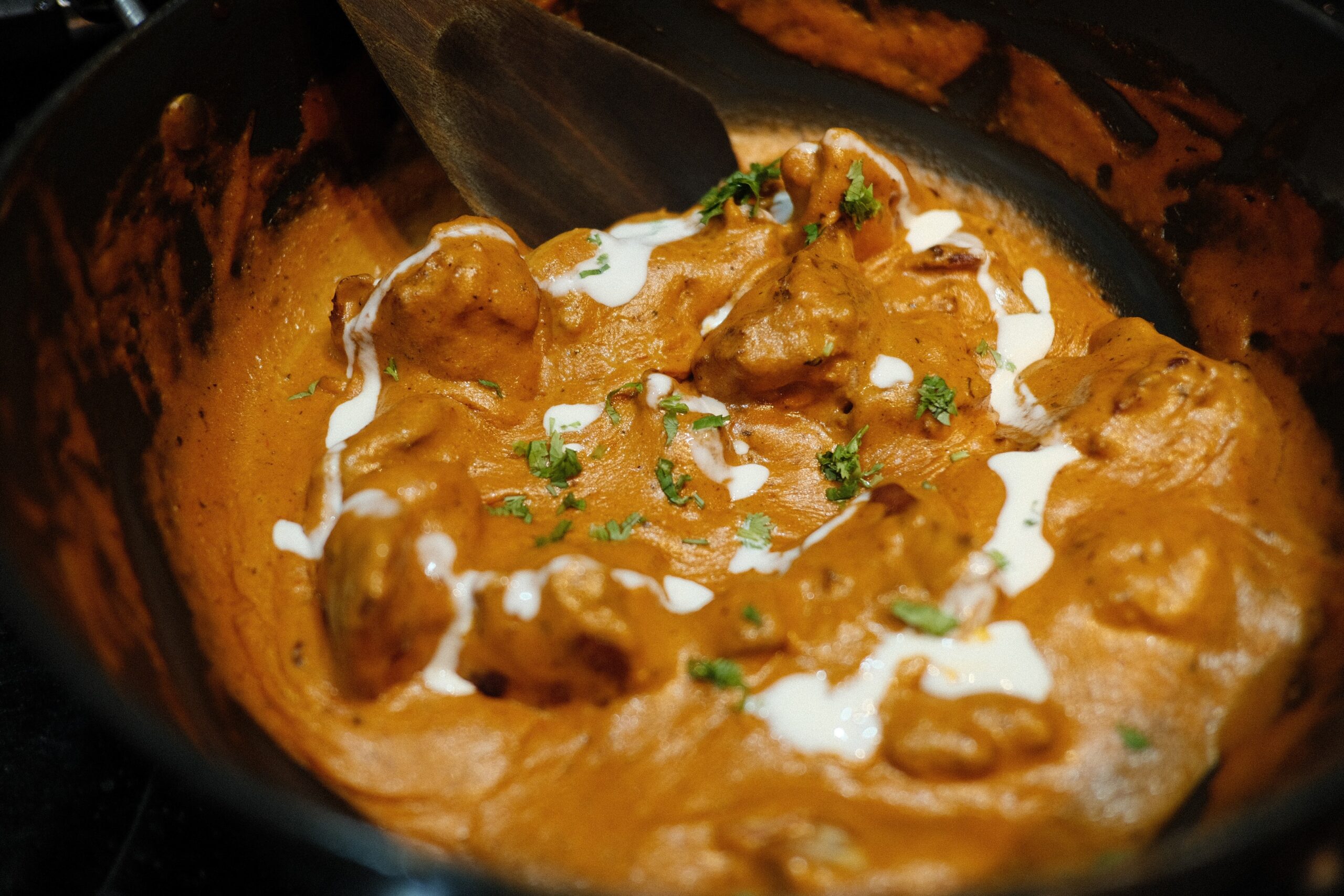 A pot of creamy butter chicken with orange sauce, garnished with swirls of cream and chopped herbs, reminiscent of the flavors you'd find at the best Indian restaurants in Leeds. A wooden spoon rests in the dish.