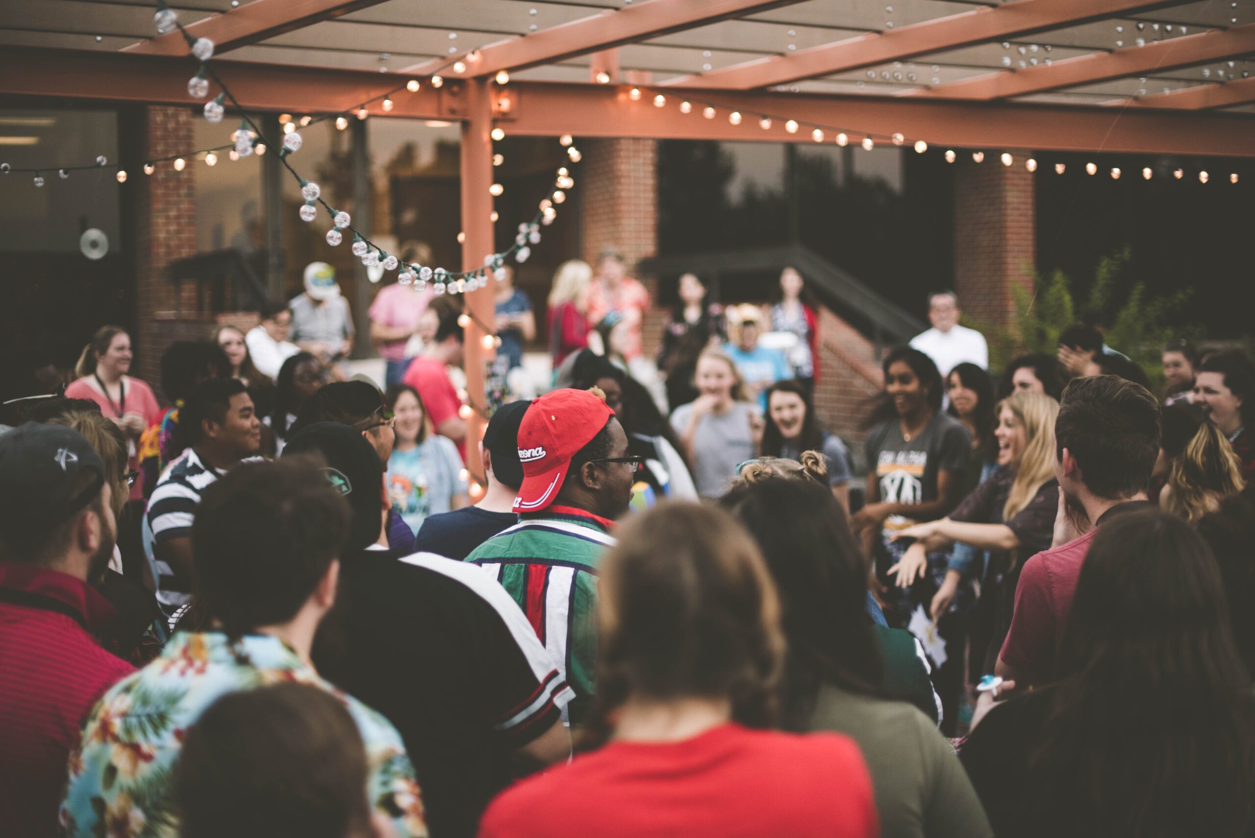 A diverse group of people, including freshers, meet in an outdoor setting under string lights. Someone in a red cap speaks in the center, surrounded by casually dressed attendees. The atmosphere is lively and social, representing various types of people coming together.