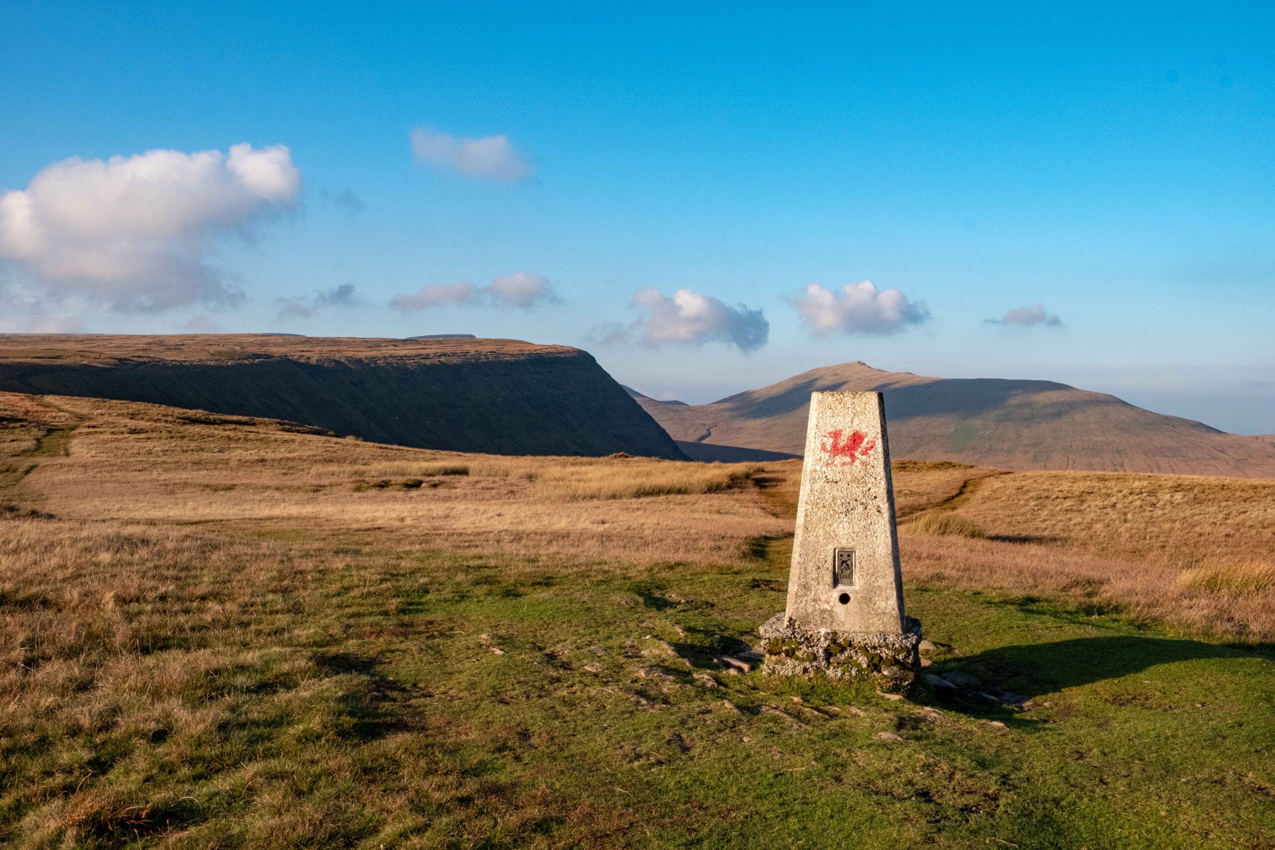 A stone pillar with a red dragon emblem stands proudly on a grassy hill near Cardiff, under a clear blue sky. Ideal for students seeking picturesque nature hikes, the scene unfolds into rolling hills stretching into the distance, dotted with scattered clouds.