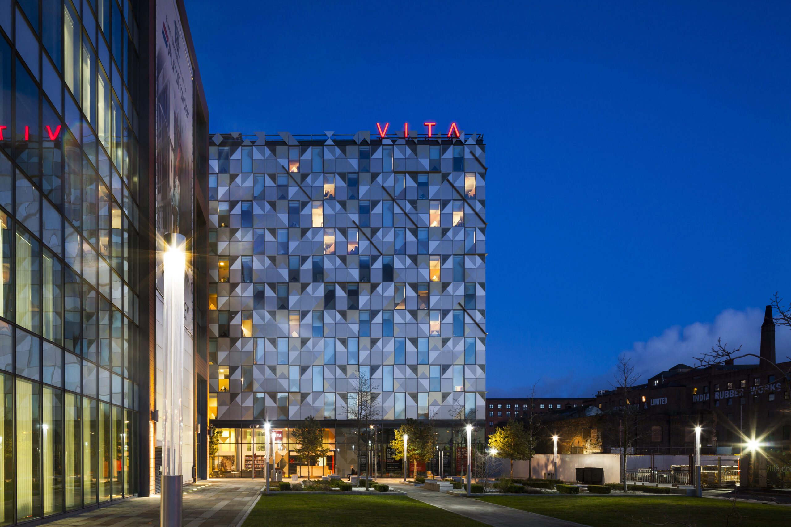 A modern building with geometric patterns is illuminated at night. The word "VITA" is displayed atop the structure. Adjacent is a glass-walled building, and a lit path with lawns stretches in front, leading to more buildings in the background.