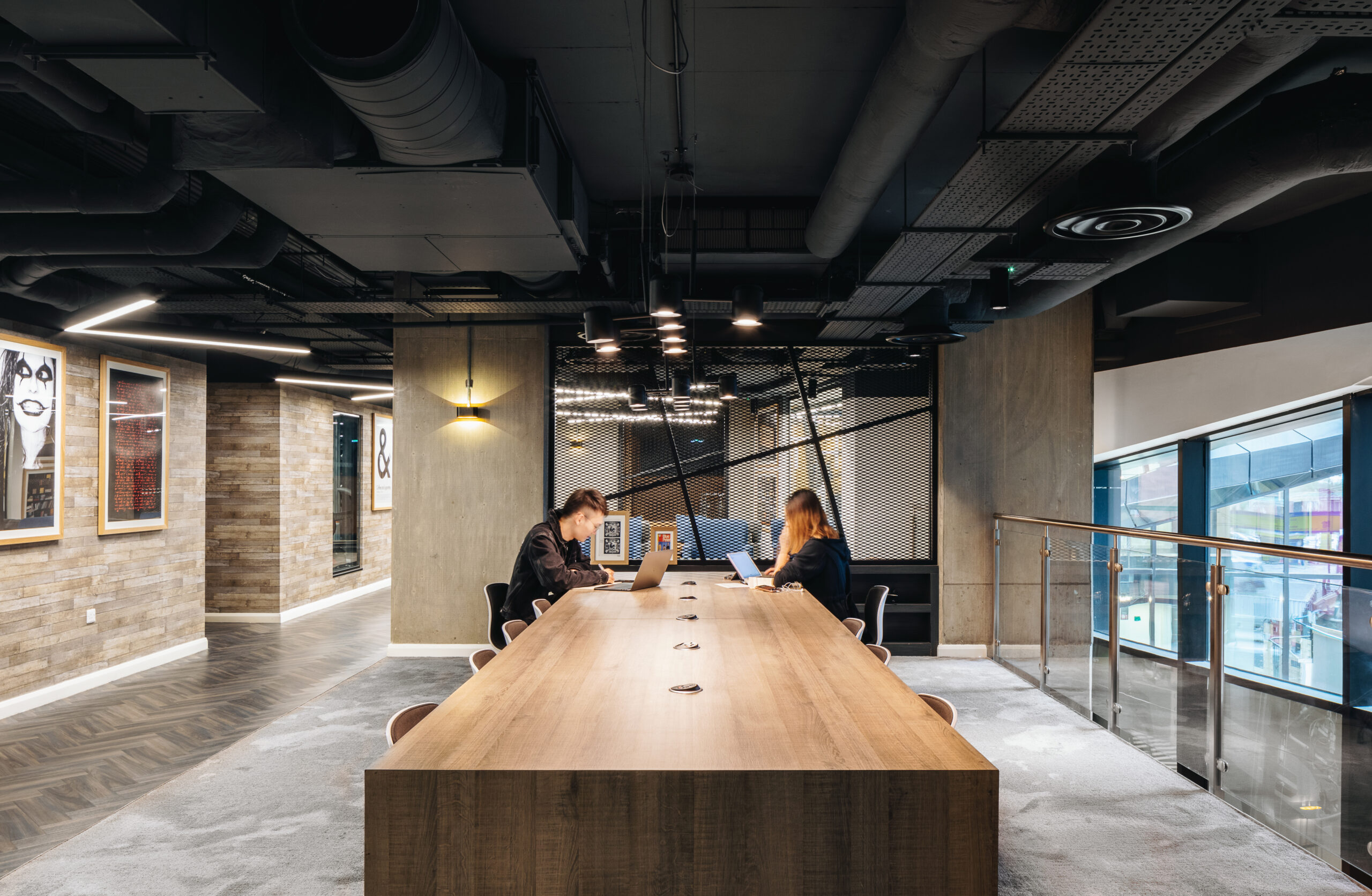 An open, modern office space with two people sitting at a long wooden table working on laptops. The room features contemporary lighting, exposed ducts, and artwork on the walls. A glass railing overlooks the area.