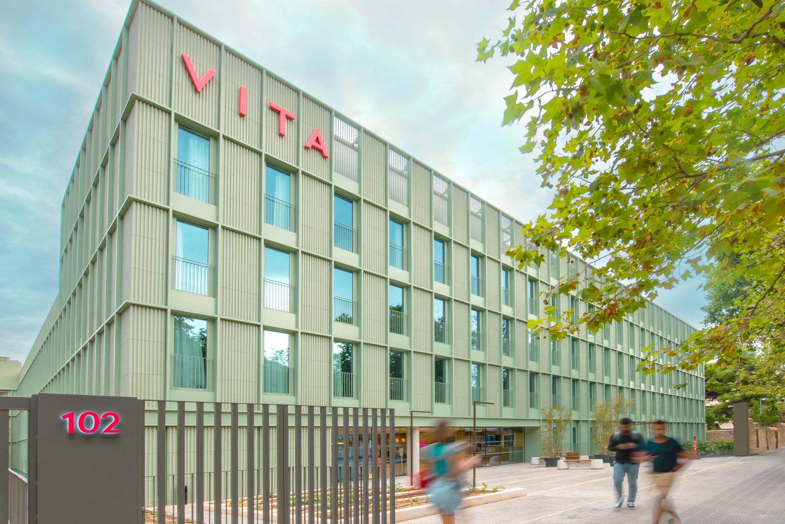 Modern building with a light green facade and large red "VITA" sign. The structure is five stories tall with numerous windows. People walk by on a path lined with trees. A sign displaying "102" is on the foreground fence.