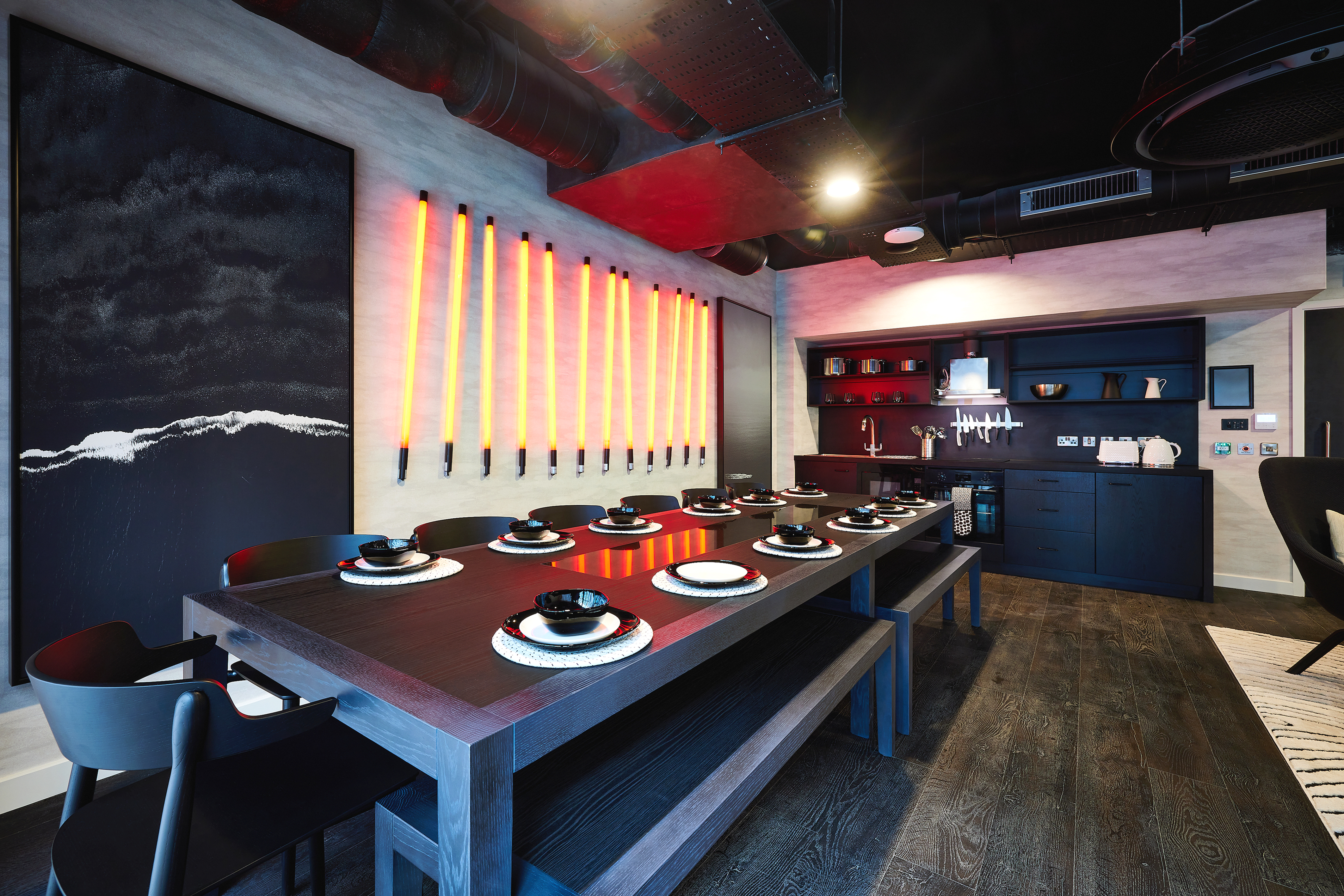 Modern dining room with a long dark wooden table set with black dishware. A large abstract painting and vertical neon lights adorn the wall. The room features dark wood flooring and exposed ceiling pipes, with a small kitchenette in the corner.