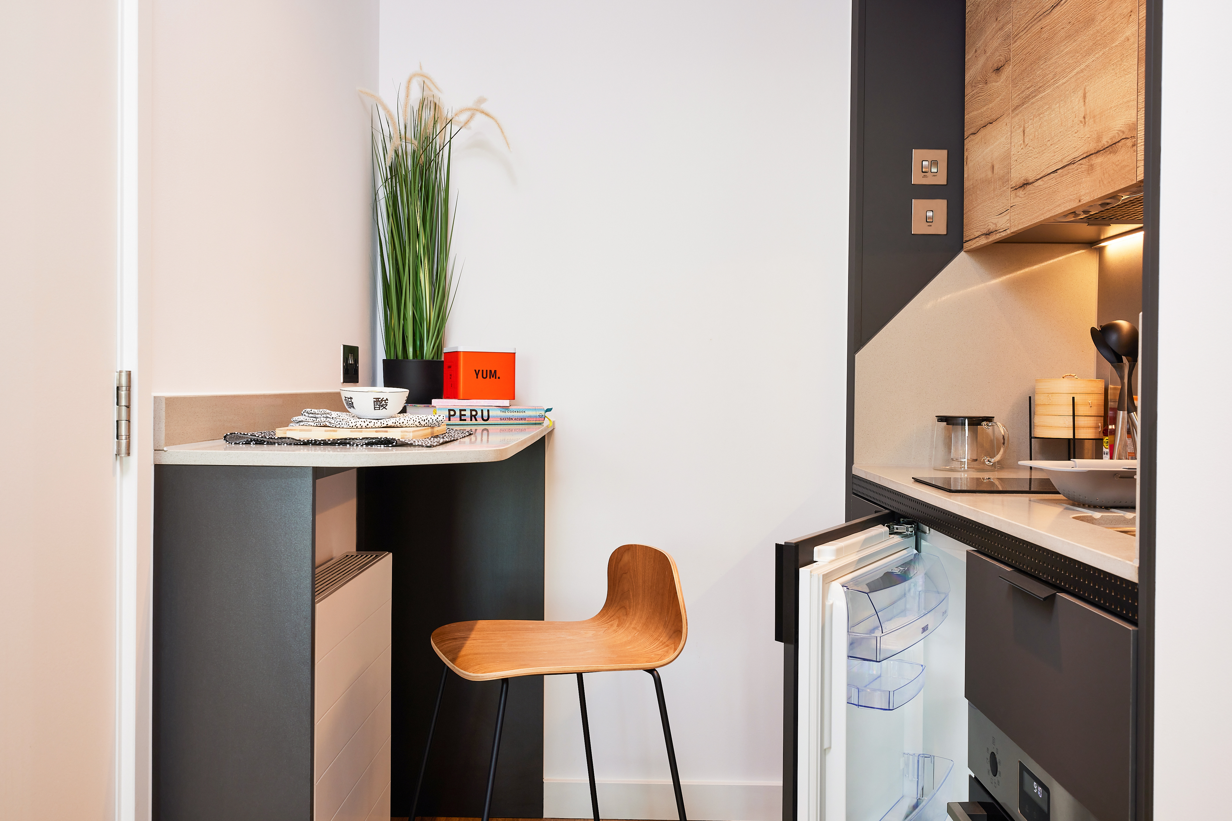 The modern kitchenette at Lewisham Exchange features a small wooden chair and a tall plant on the counter. An orange book titled "YUL" sits beside a bowl and newspapers. This compact kitchen area includes a stove and an open mini fridge, set against white and grey walls.