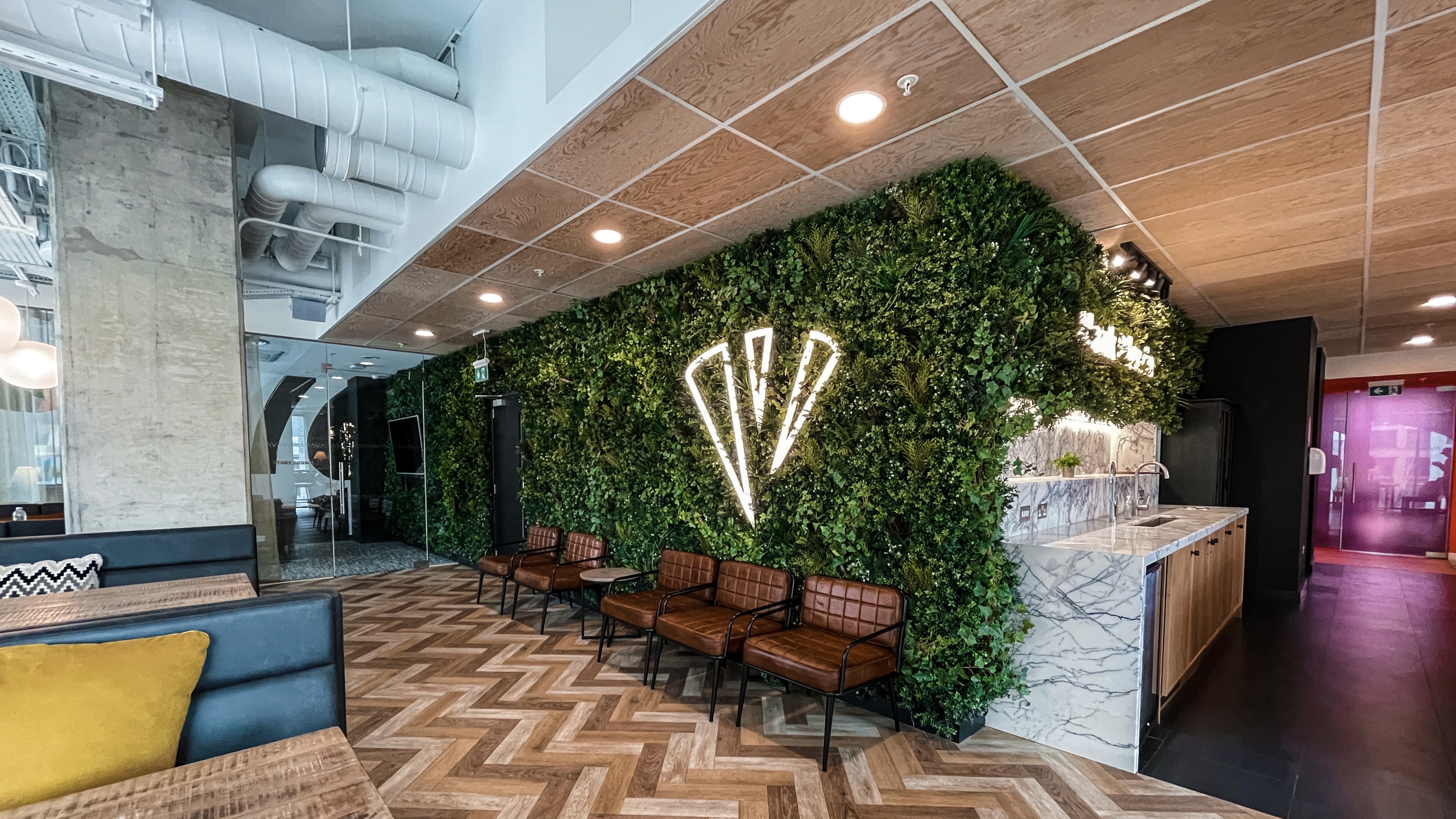 Modern office lounge in Lewisham Exchange with a green plant wall featuring illuminated geometric shapes. Brown leather chairs line the wall, while chevron-patterned wooden floors and tables with mustard-yellow cushions create a cozy atmosphere. Ceiling features visible ducts.