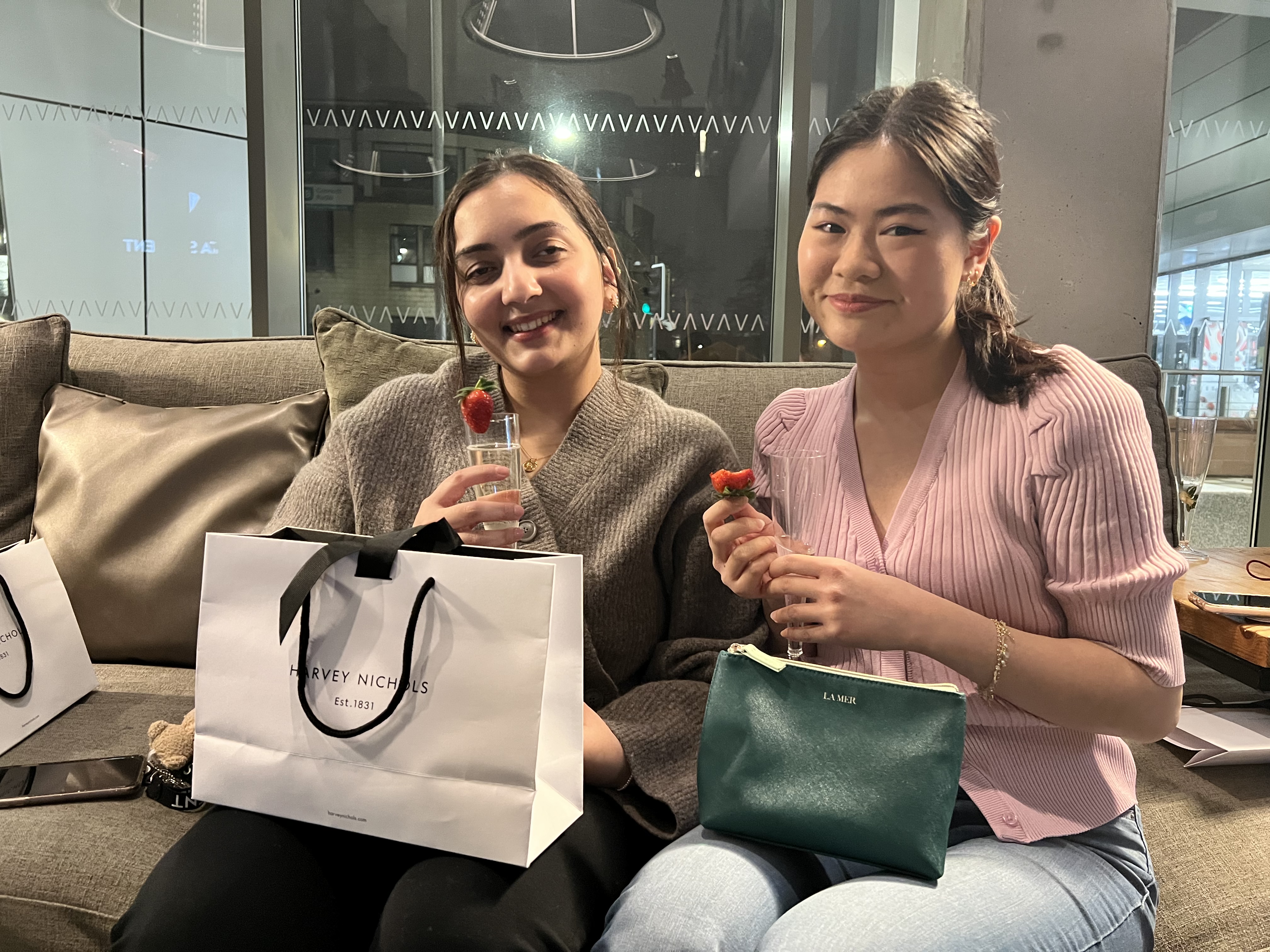 Two women sit on a sofa, smiling and holding glasses with strawberries. A white shopping bag marked "CN" is beside one, while the other has a green purse. They're in a brightly lit setting with large windows, reminiscent of a chic urban General UK atmosphere.