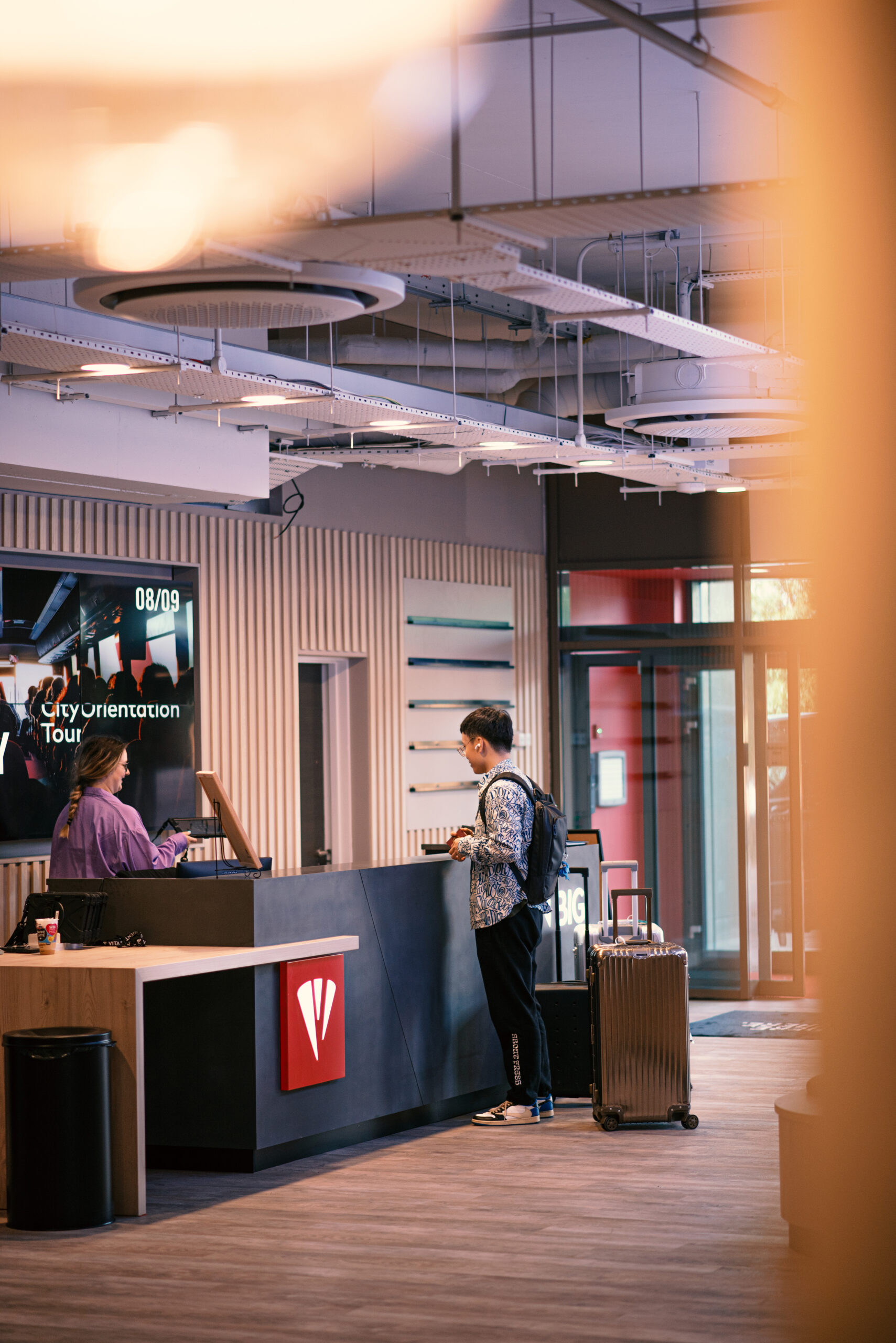 An international student wearing a backpack stands at a modern reception desk in an open lobby area with a suitcase. A screen behind the desk displays "City Orientation Tour." The setting has industrial-style decor and large windows, welcoming newcomers from around the world.
