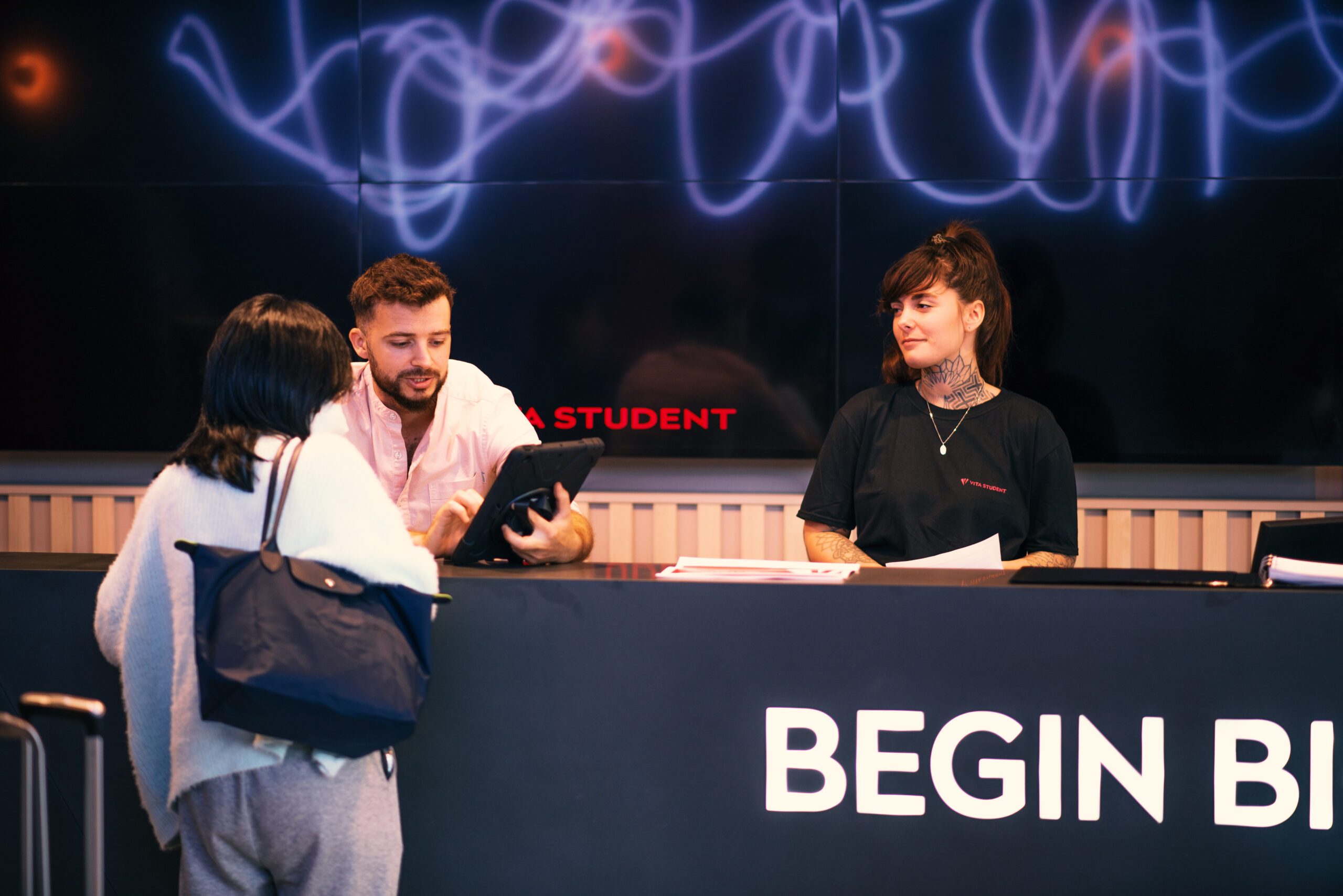 A woman stands at a counter talking to two staff members. The man, holding a tablet, and the tattooed woman beside him share warm smiles. The counter displays "BEGIN BIG," embodying the vibrant essence of vita. Neon-style writing glows on the wall behind them, breathing life into the scene.