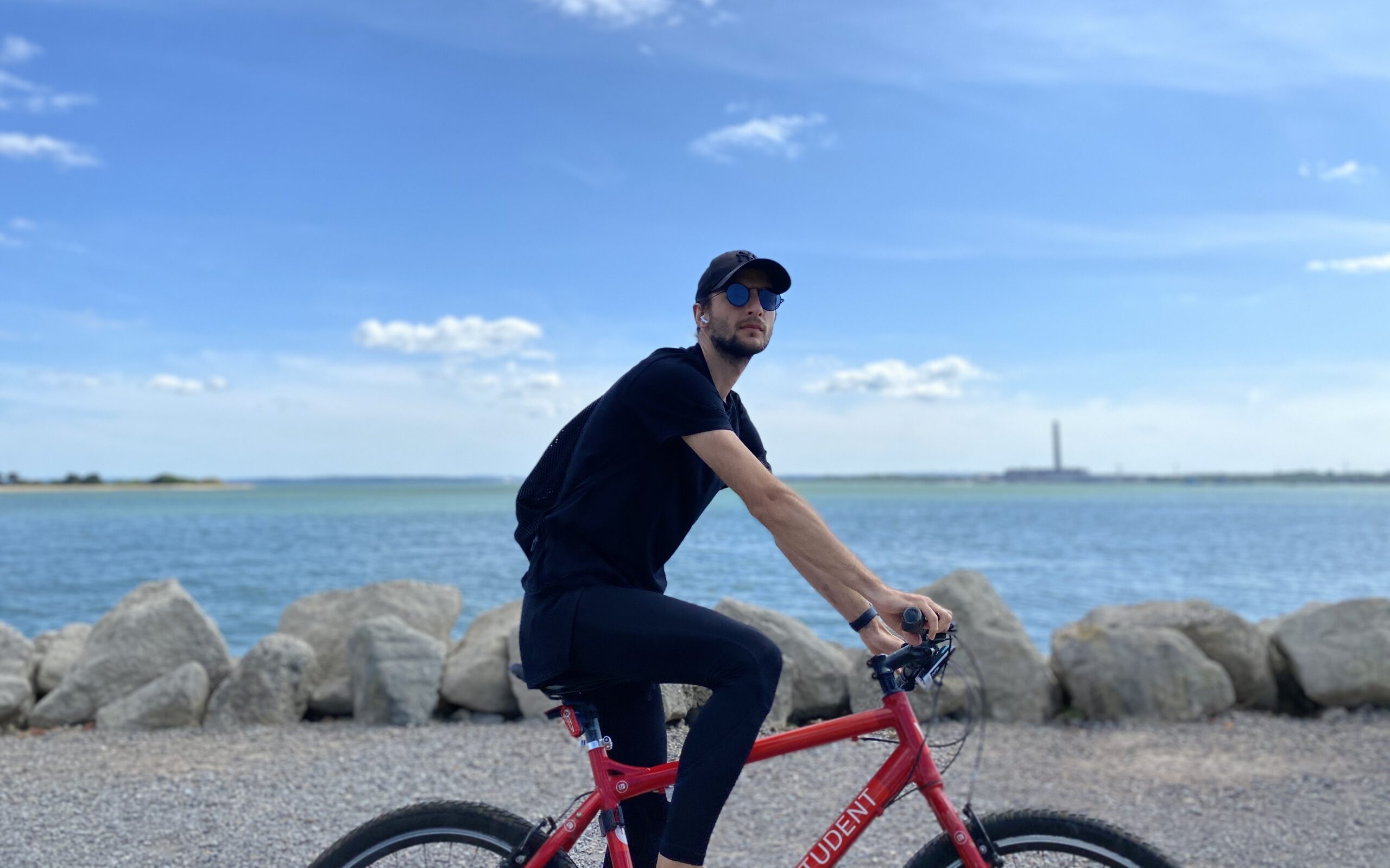 A person in a black outfit and cap rides a red bicycle along a rocky shoreline, perhaps taking a break from student accommodation. The sky is clear with a few clouds, and water stretches into the distance.