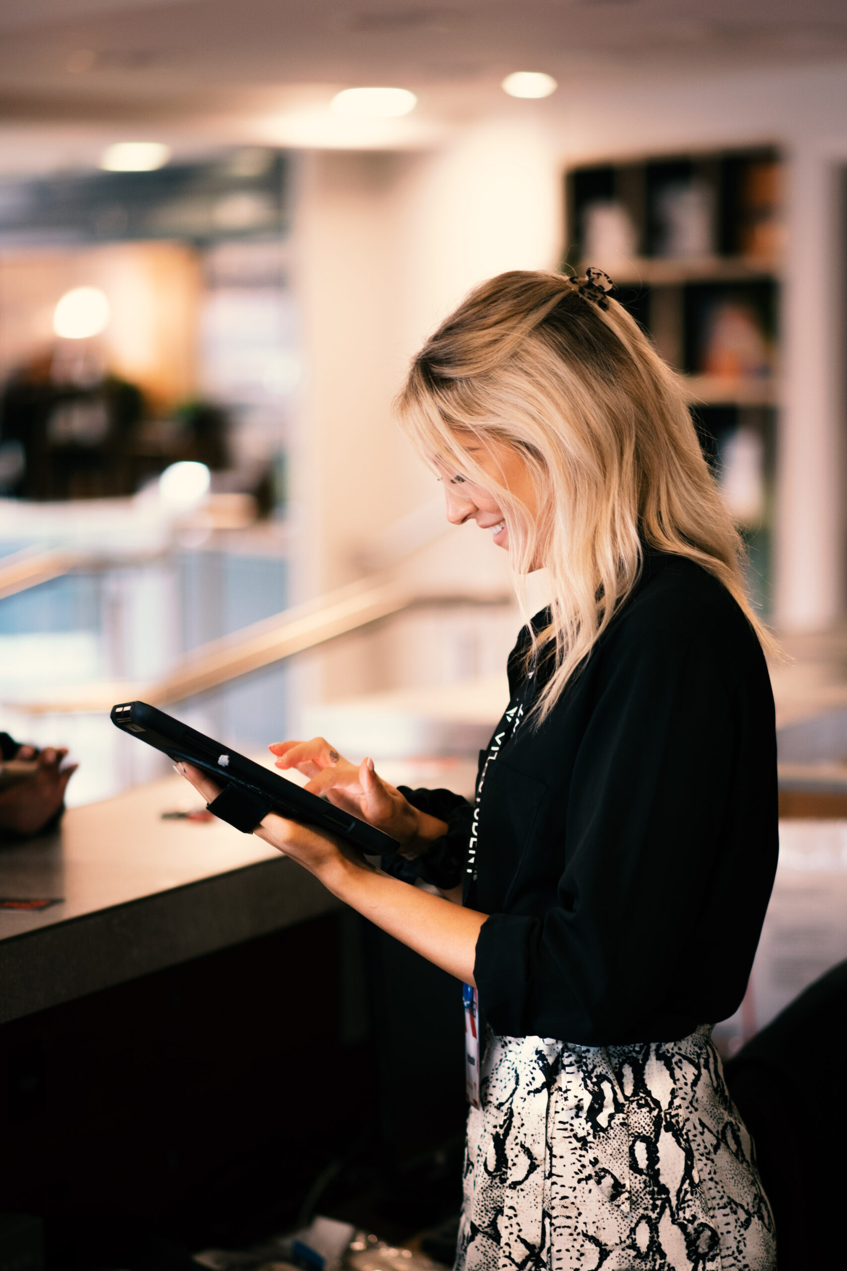 A woman with long blonde hair, wearing a black top and a white patterned skirt, stands at a counter using a tablet for parenting advice. The background is softly focused with shelves and lights.