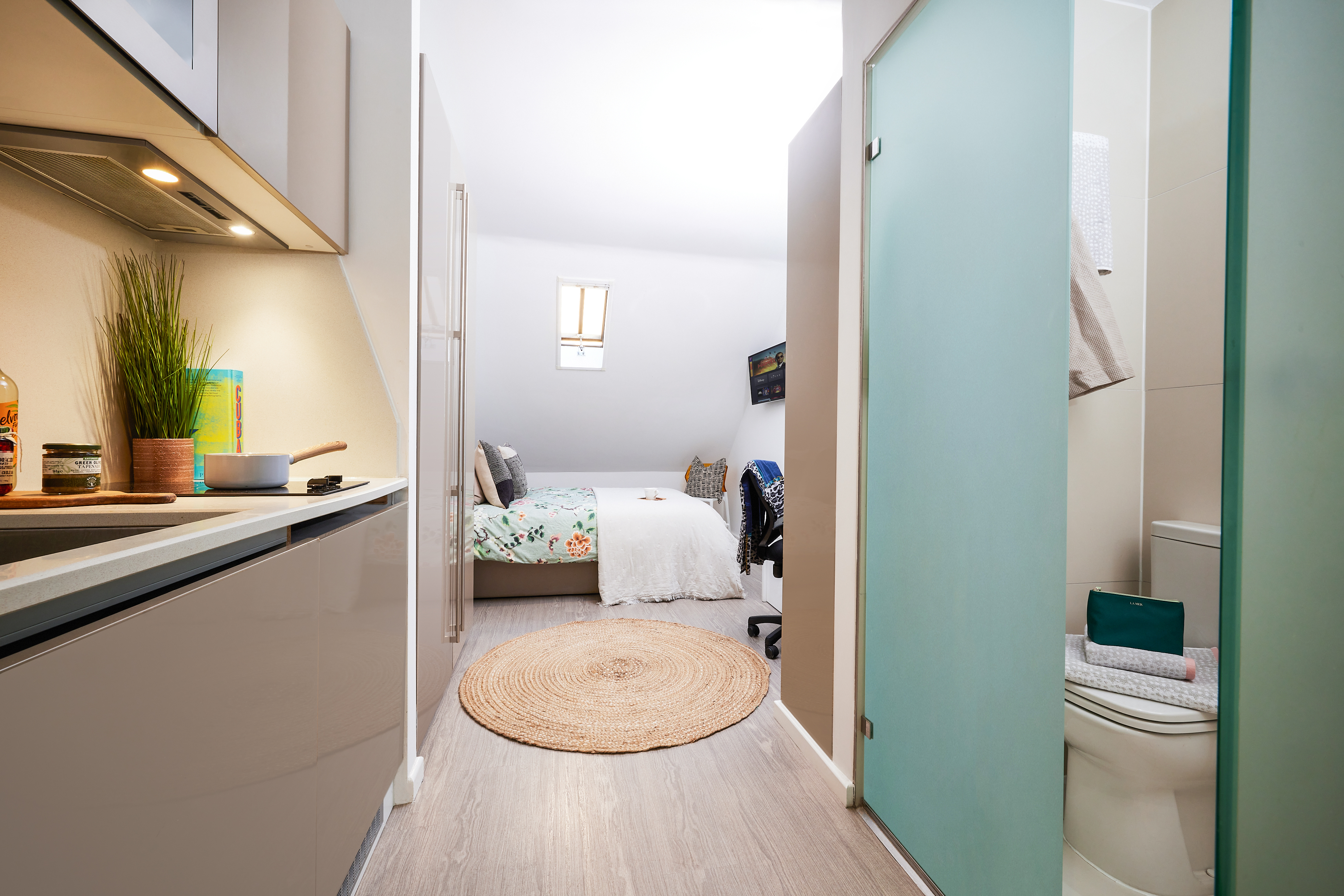 A compact studio apartment on Crosshall Street features a kitchenette on the left, a bathroom with a frosted sliding door on the right, and a cozy bedroom adorned with a floral bedspread at the end. A round woven rug graces the light wood floor while a small window invites natural light.