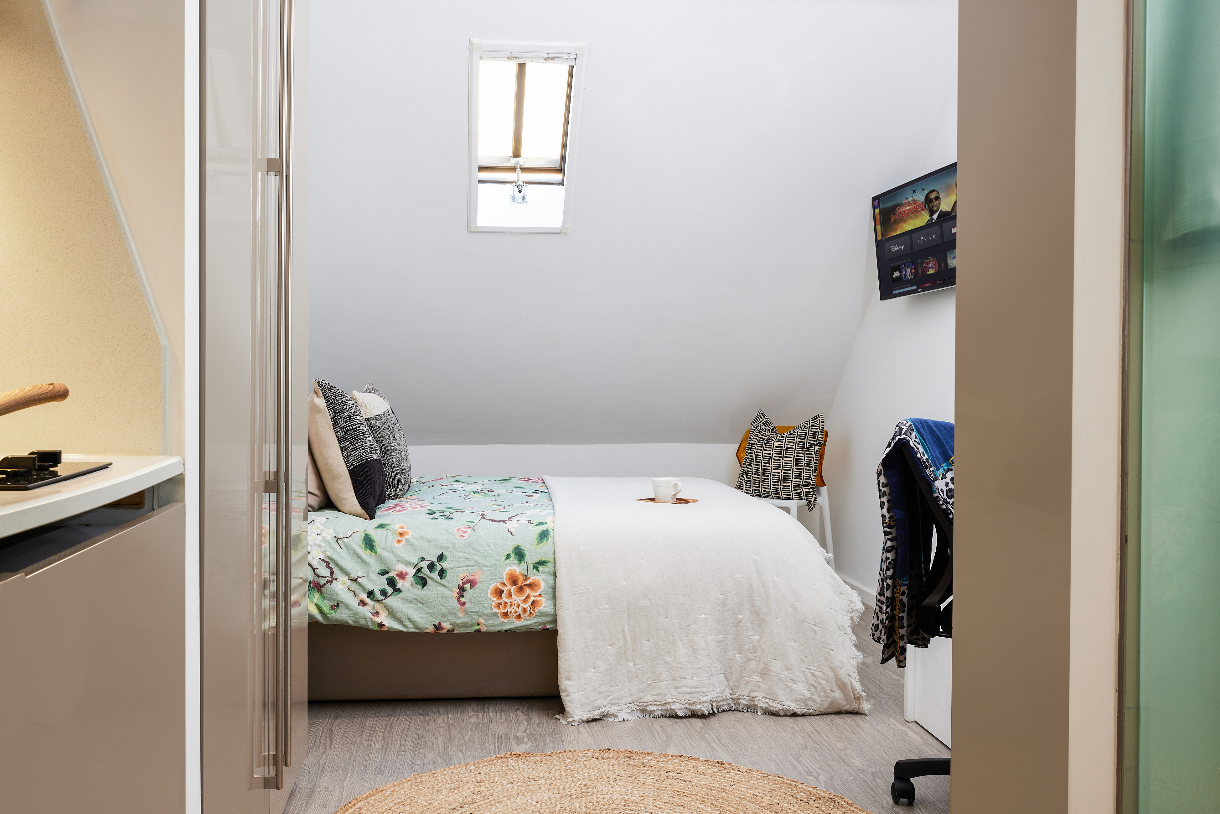A cozy attic bedroom on Crosshall St. features a sloped ceiling and a single bed with floral bedding. A small desk holds a computer screen, while a round woven rug adorns the floor. The skylight window above casts natural light into this charming space.