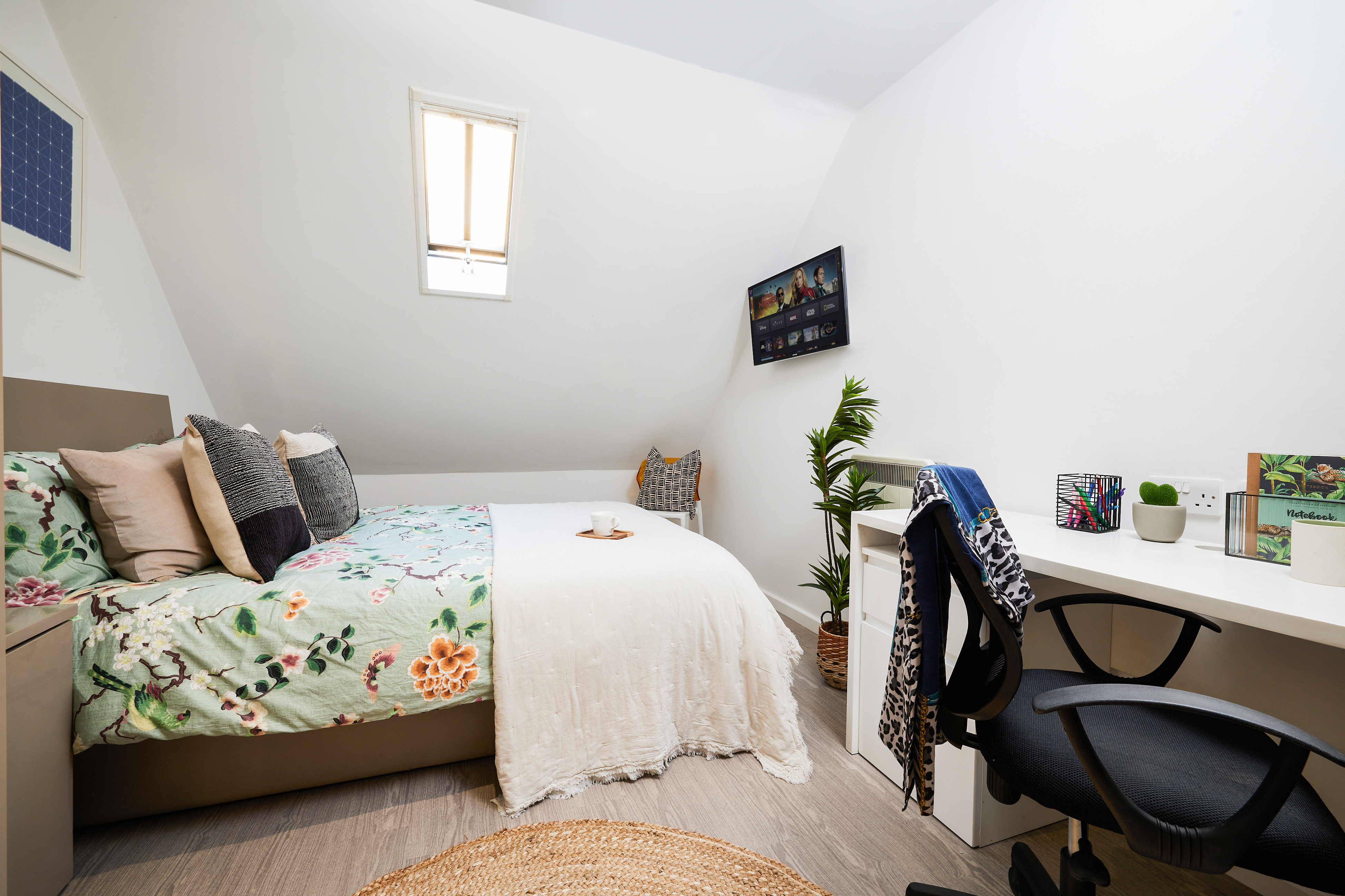 Nestled under a sloped ceiling, this cozy Crosshall St. bedroom showcases floral bedding, a small window, and a wall-mounted TV. A desk with an office chair complements the space, while a potted plant and woven rug enhance its warm ambiance.