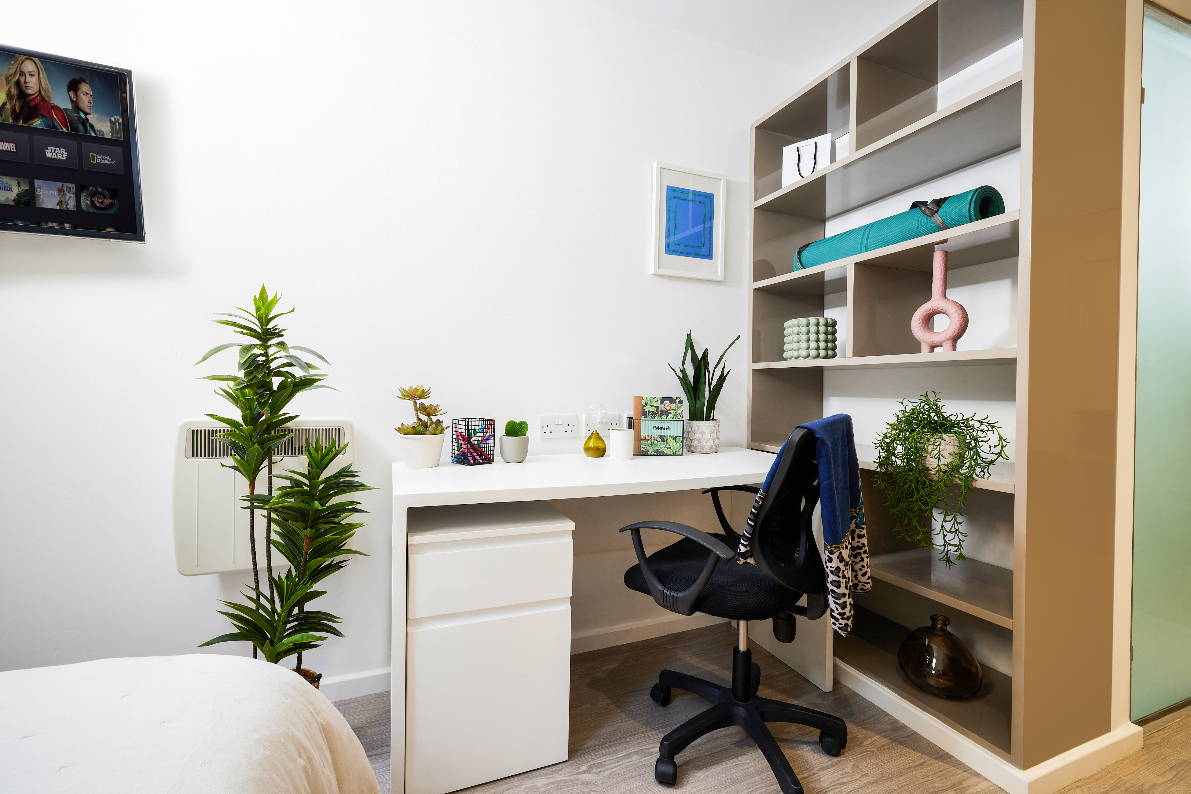 Nestled in a chic Crosshall St. locale, this modern study space features a white desk paired with a black swivel chair. The desk hosts plants, a mug, and accessories. To the right is a beige shelving unit adorned with decor and a rolled-up yoga mat, alongside a wall-mounted TV and lush greenery.