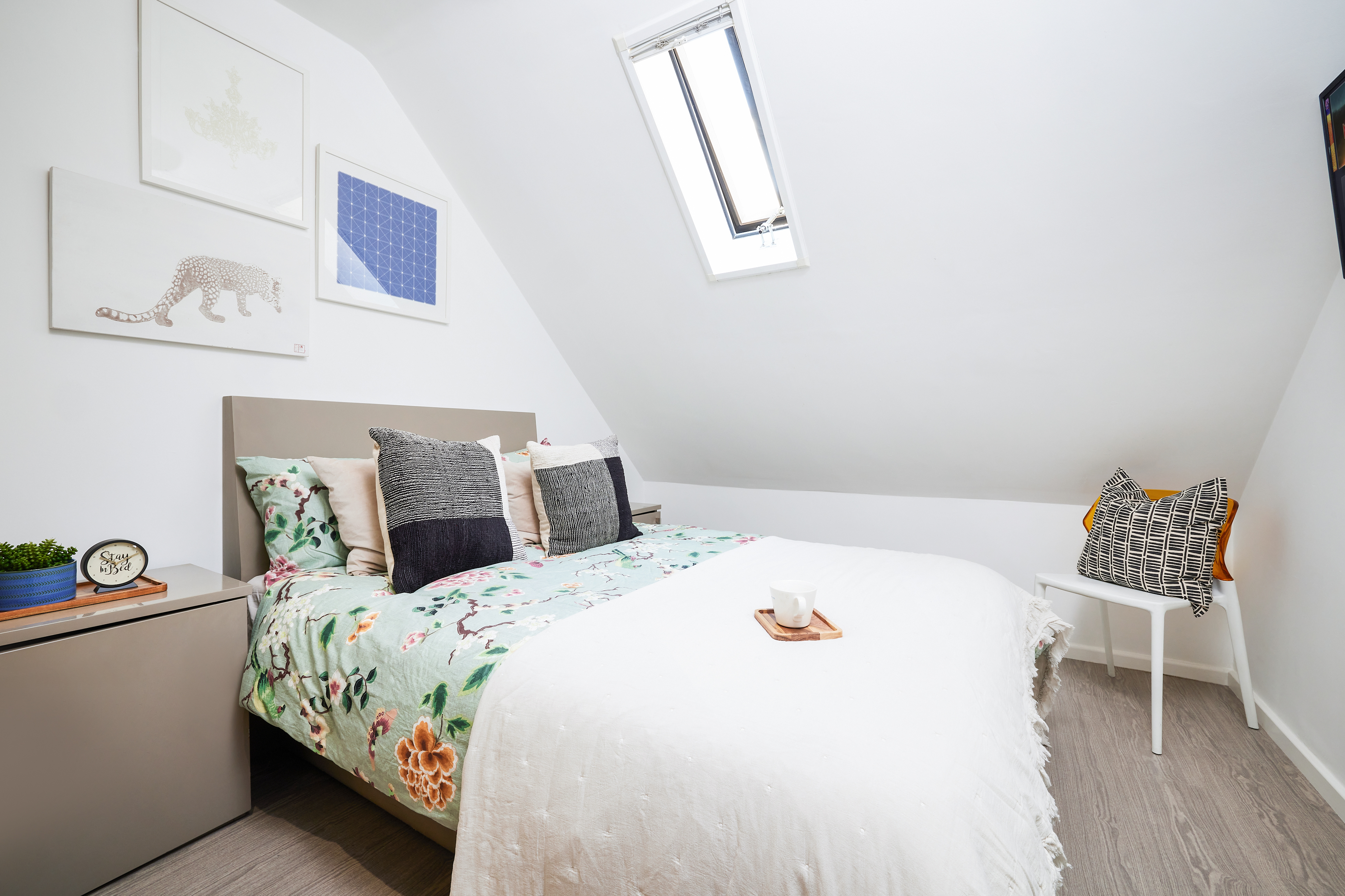 Crosshall St. offers a cozy attic bedroom with a slanted ceiling, featuring a bed adorned with floral bedding and decorative pillows. A small window allows natural light to illuminate the space, which also includes framed artwork and a small white side table topped with a patterned pillow.