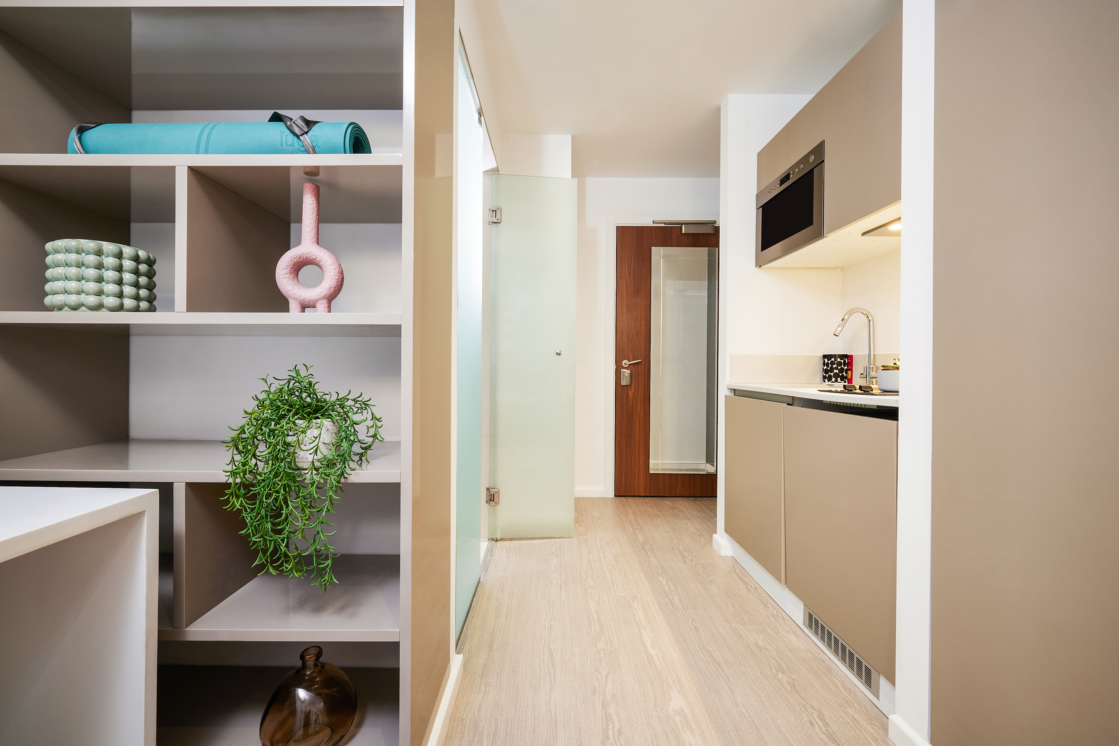 A modern, compact kitchen on Crosshall St. features beige cabinets and a small countertop. On the left, open shelves display decorative items, like a rolled yoga mat and plant. A wooden door stands at the back, while natural light floods the space, creating an inviting atmosphere.