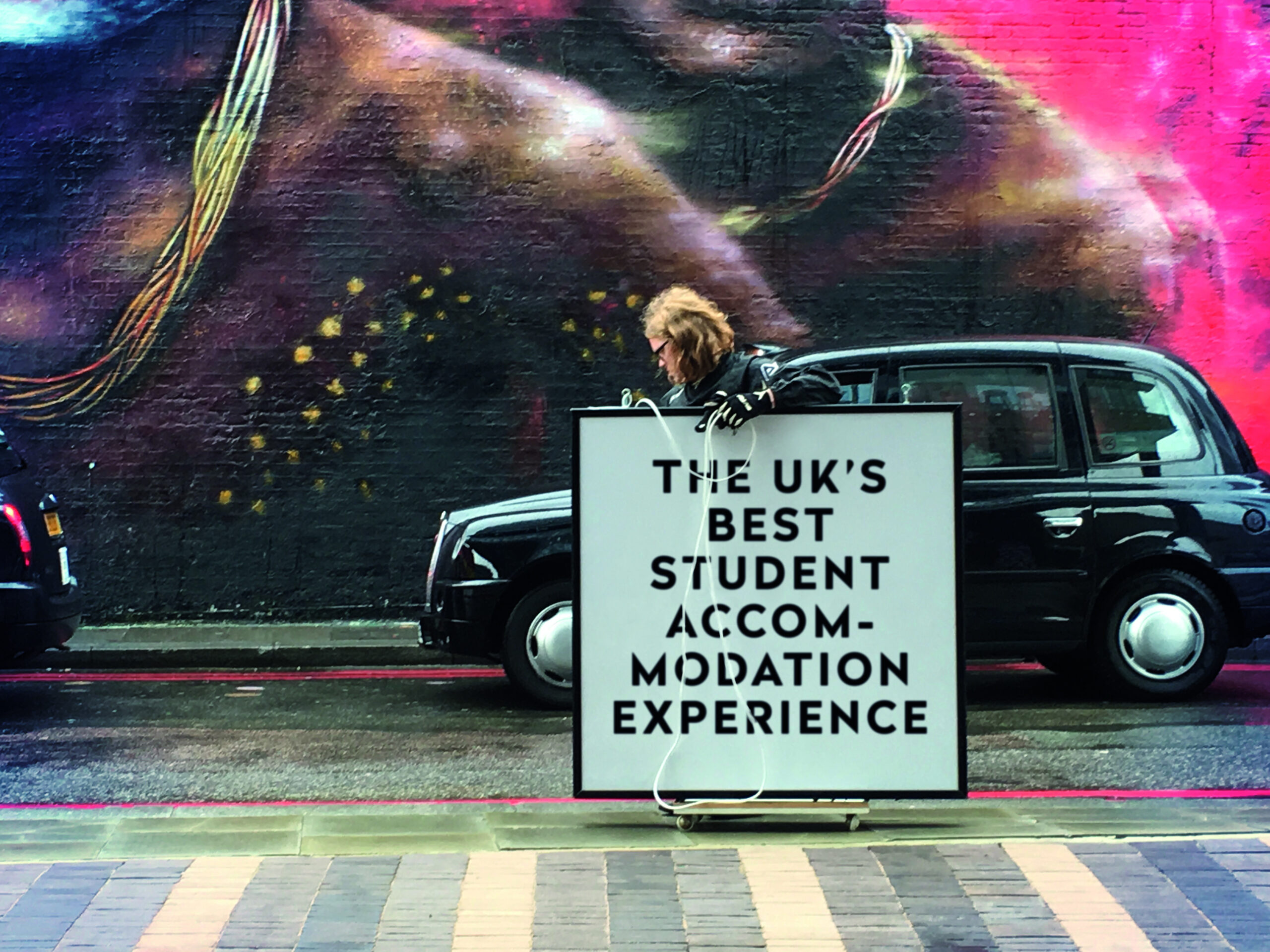 A person stands on the side of the road next to a black taxi, holding a large sign that reads, "The UK's Best Student Accommodation Experience for International Students." A colorful mural is visible on the brick wall behind them.