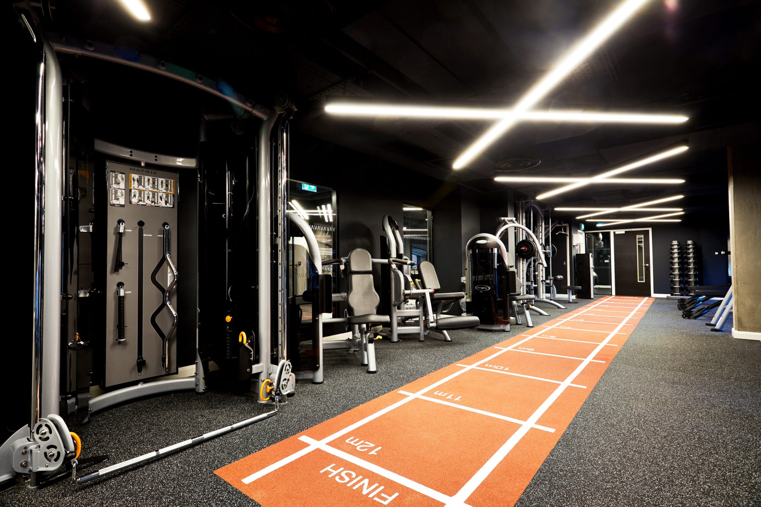 A modern gym with various exercise equipment, including machines and weights, along the sides. An orange athletic track runs across the floor with marked distances. The ceiling features sleek, crisscrossing lights creating a dynamic atmosphere.