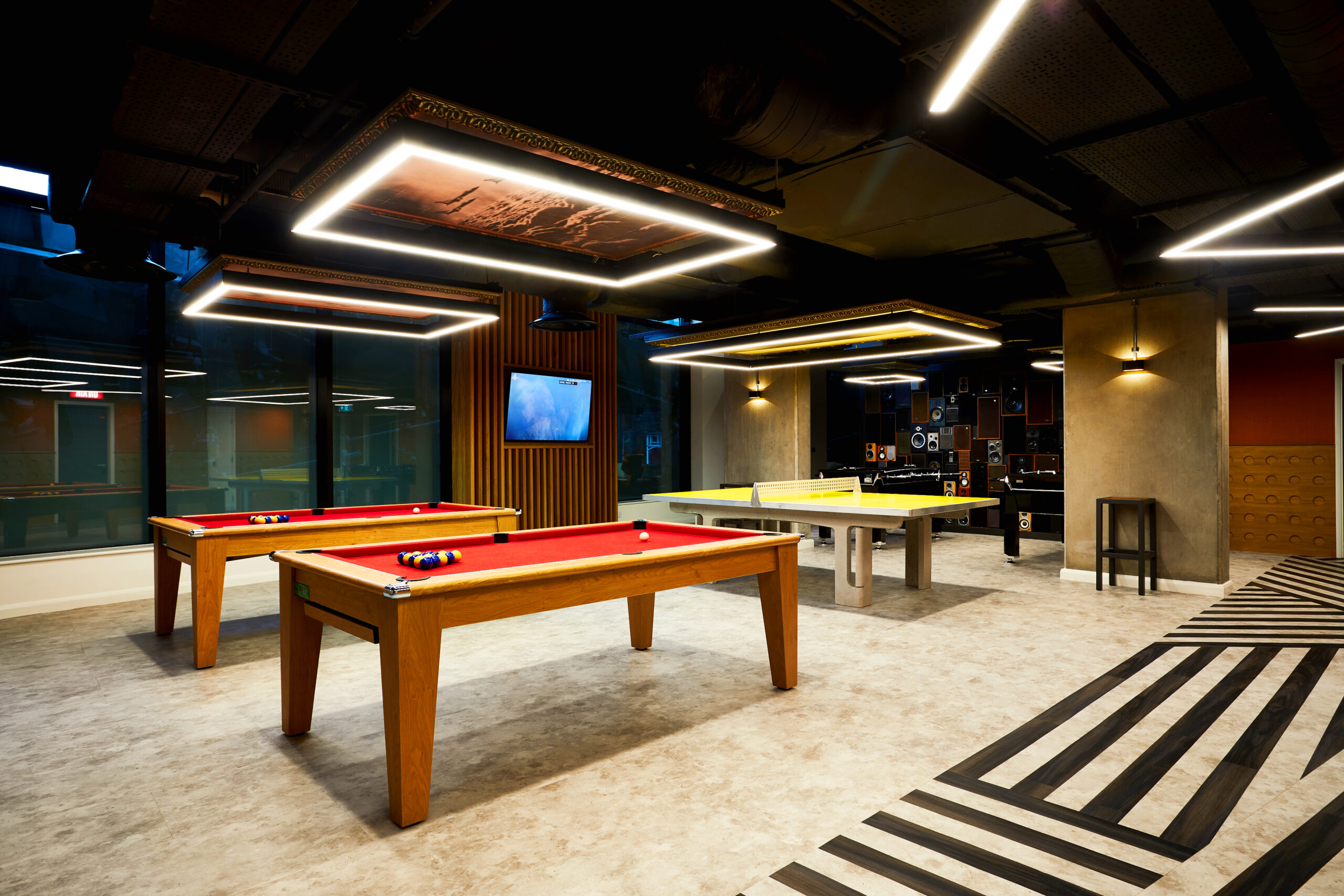 A modern game room with three pool tables featuring red and yellow felt. Overhead lighting includes rectangular fixtures. The room has dark ceilings, a wall-mounted TV, and a striped pattern on the floor.