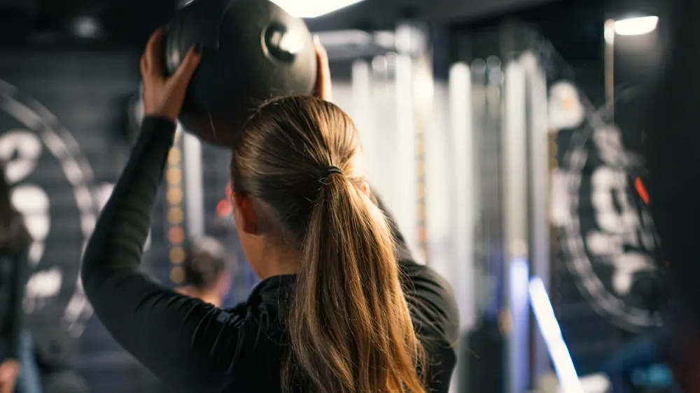 A person with long hair in a ponytail holds a medicine ball above their head in a gym, embodying the latest fitness trends. The background is blurred, featuring gym equipment and another individual. The scene highlights physical activity and focus, reminiscent of popular TikTok fitness challenges.